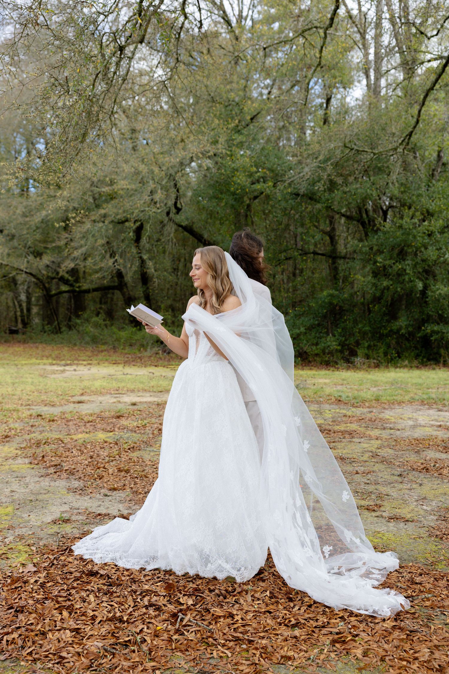 Bride reads vow book while standing back-to-back with groom under moss-draped trees outdoors.