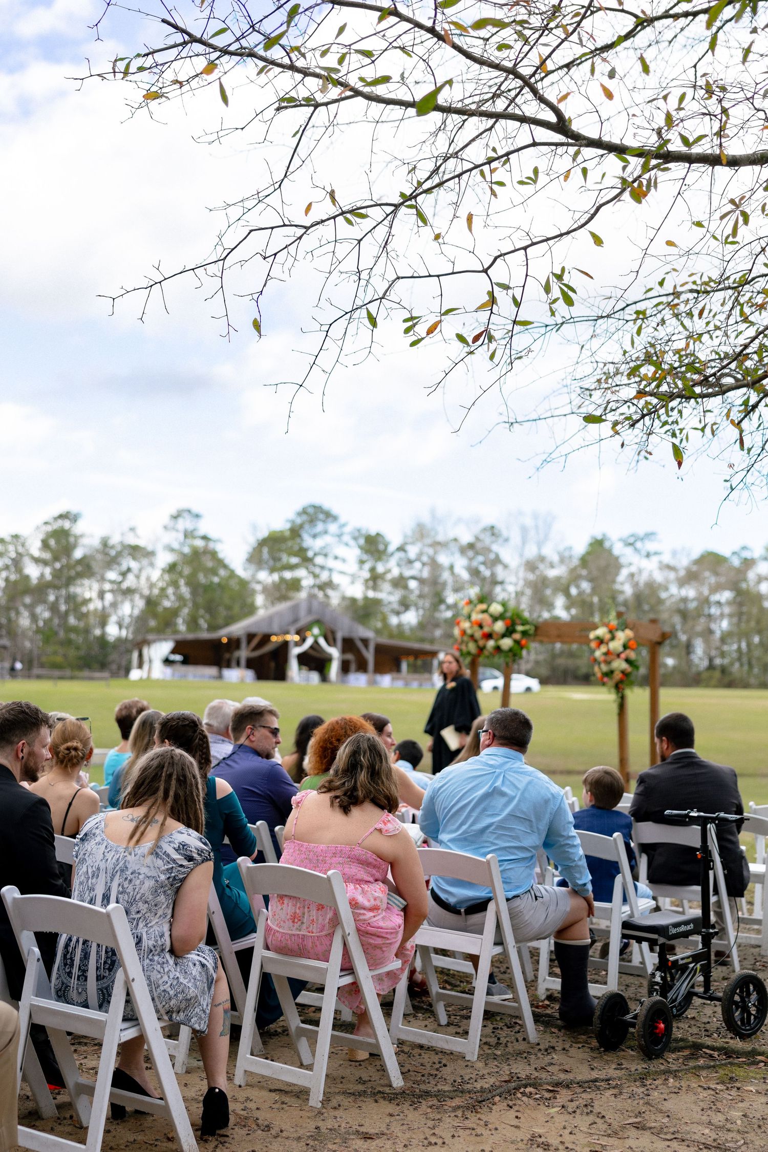 Guests seated in white chairs at an outdoor wedding ceremony with a floral arch altar in the background.