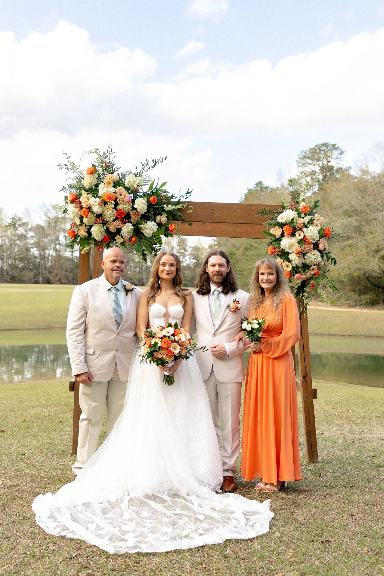 Wedding party of four poses before wooden arch with orange floral arrangements beside a pond.