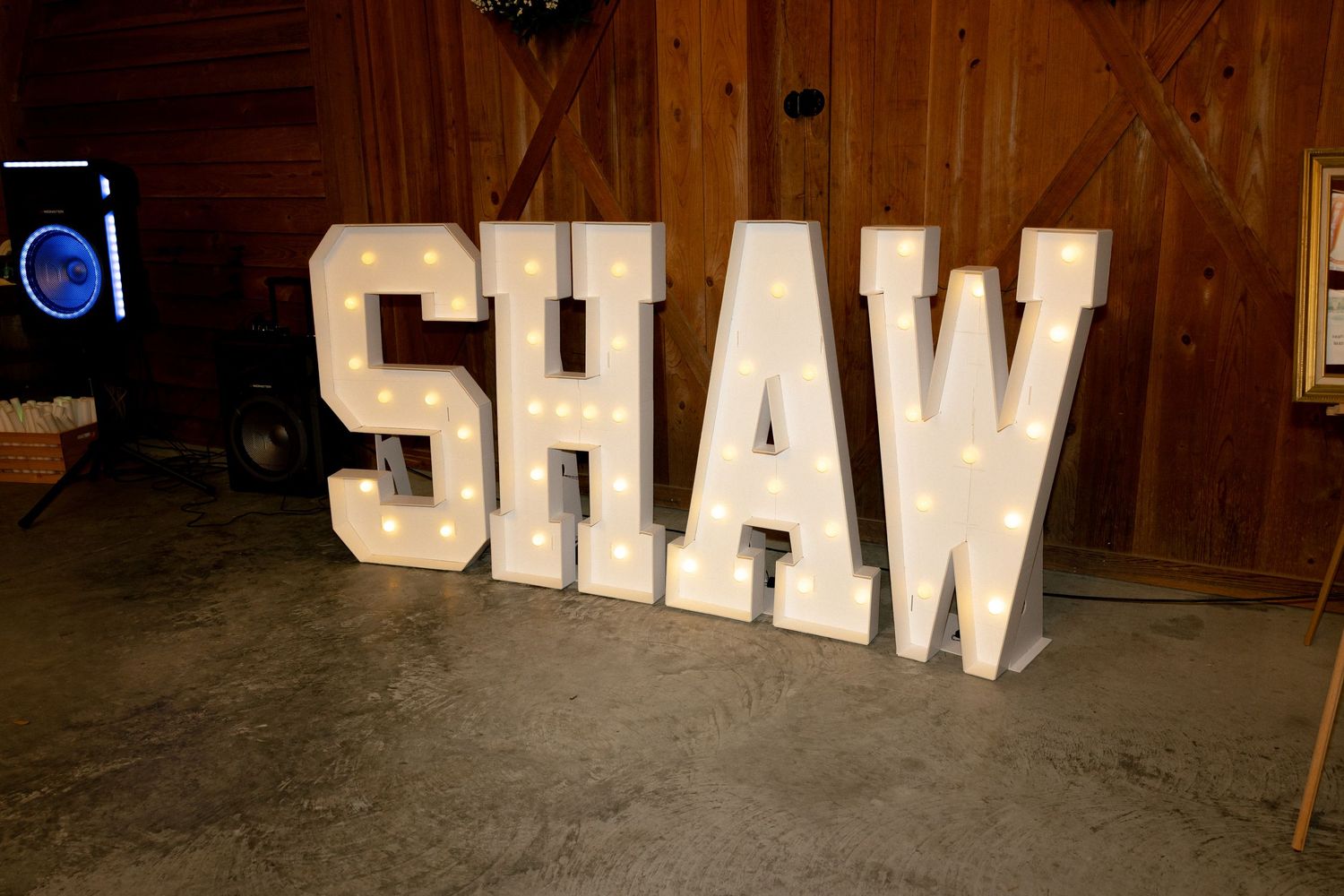 Illuminated marquee letters spelling SHAW displayed on the floor of a rustic barn wedding venue.