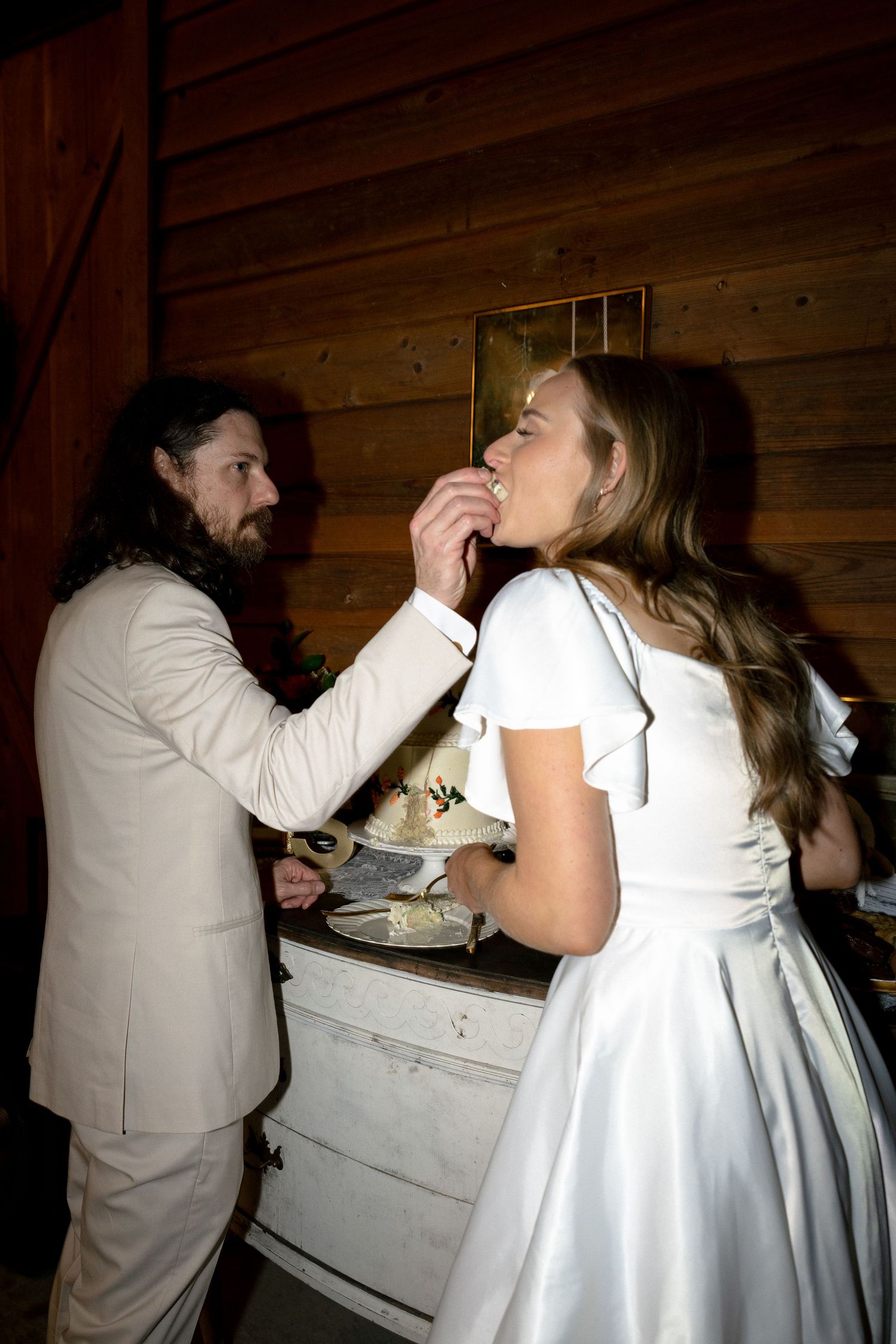Groom in beige suit feeds wedding cake to bride in white dress at rustic barn reception.