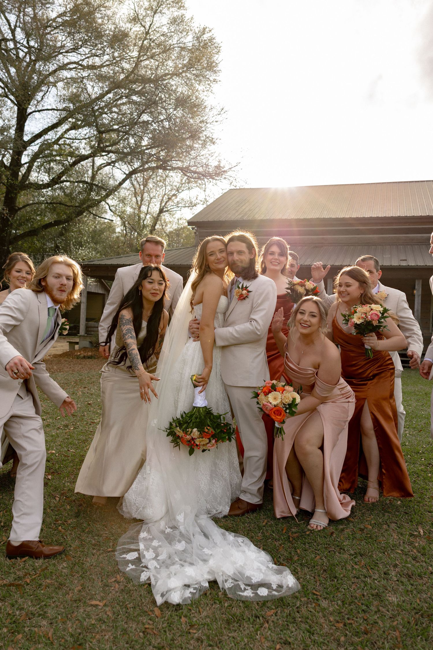 Bride and groom hug while cheerful wedding party poses playfully around them in warm outdoor sunlight.