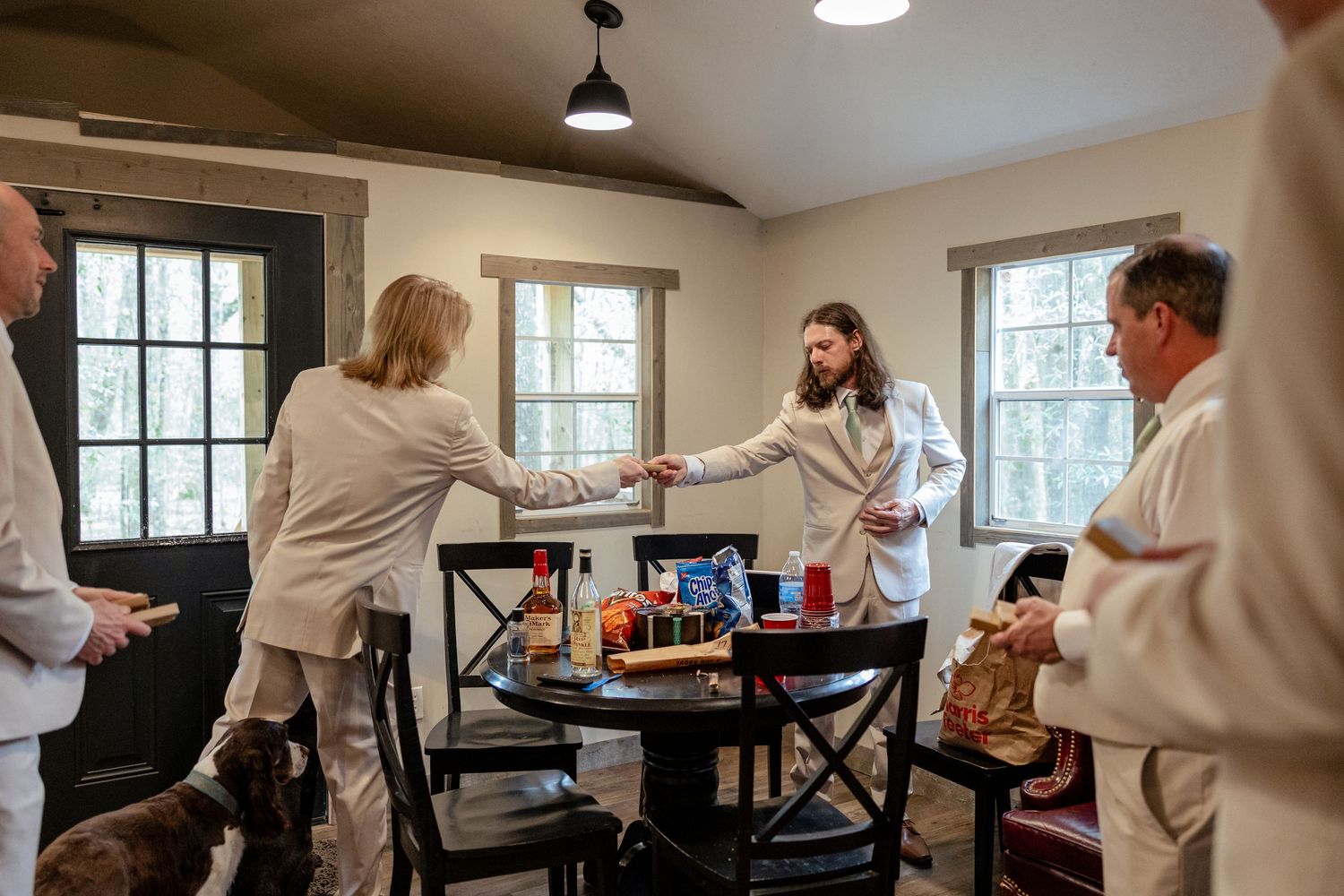 Wedding party in white suits gathers around a table sharing gifts in a rustic venue room.
