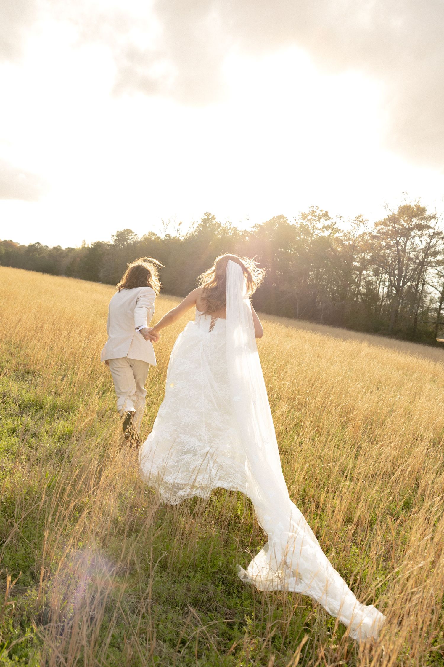 Bride and groom running hand in hand through a golden sunlit field, wedding dress and veil flowing in the breeze.