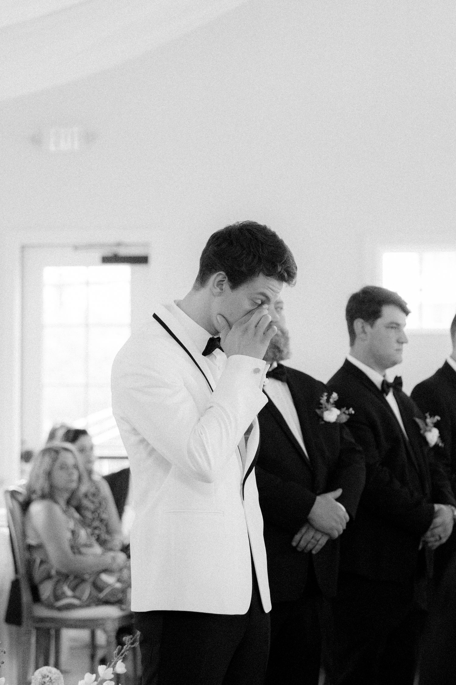 Emotional groom in white tuxedo jacket wiping tears at wedding ceremony with groomsmen standing behind him.