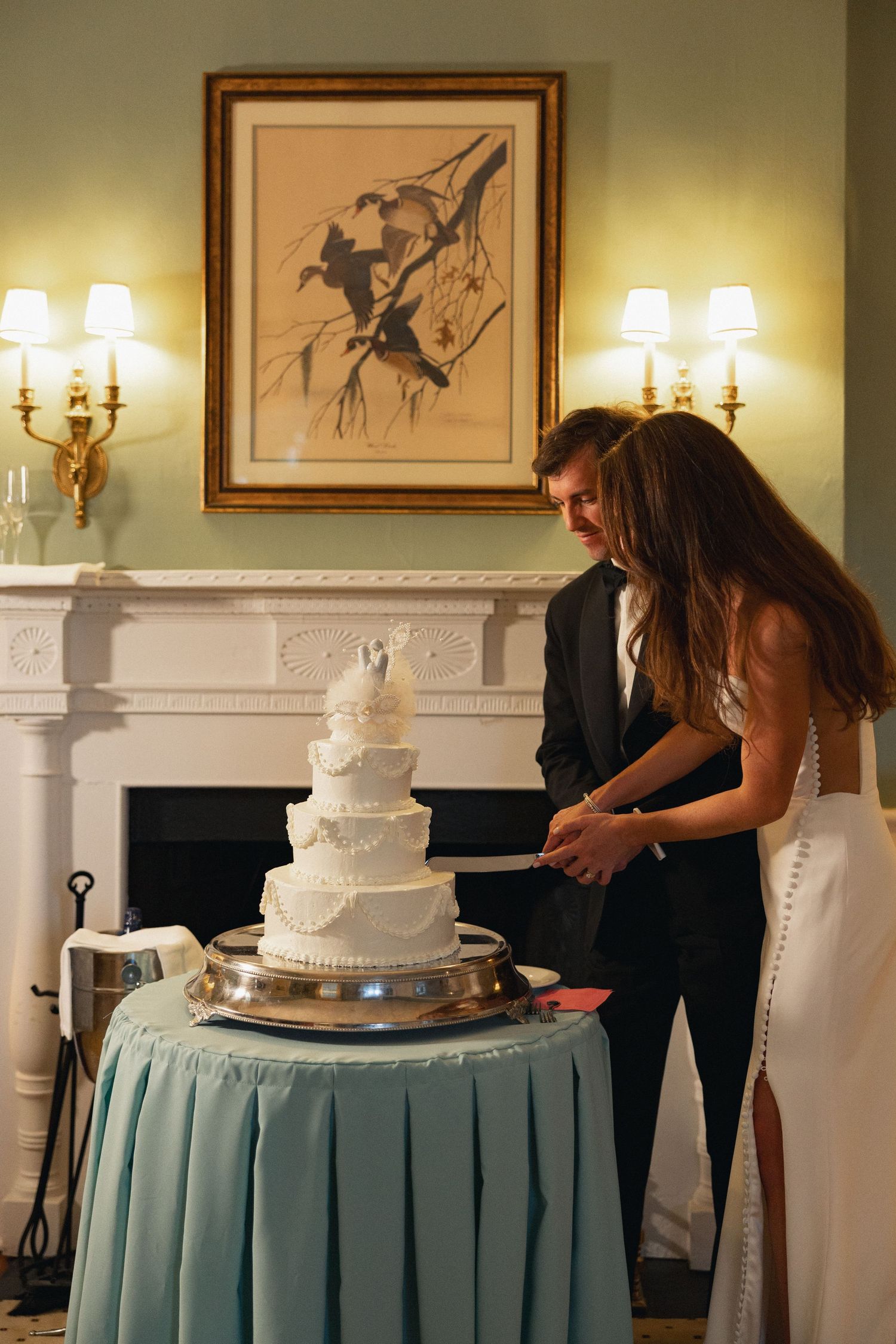 Bride and groom cutting a white tiered wedding cake on a teal draped table by an elegant fireplace.