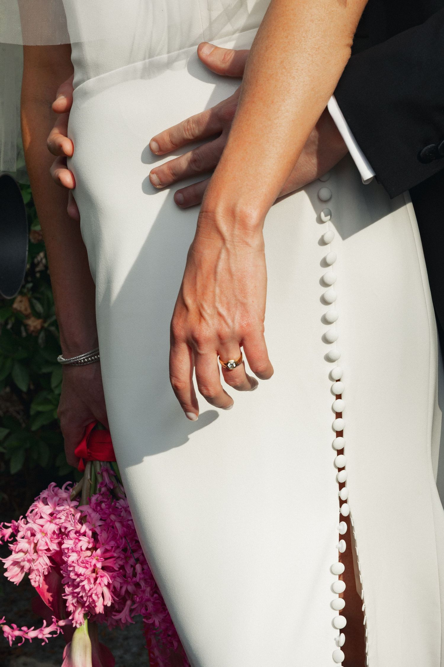 Close-up of bride and groom hands with wedding ring on white dress with button detail and pink flowers.