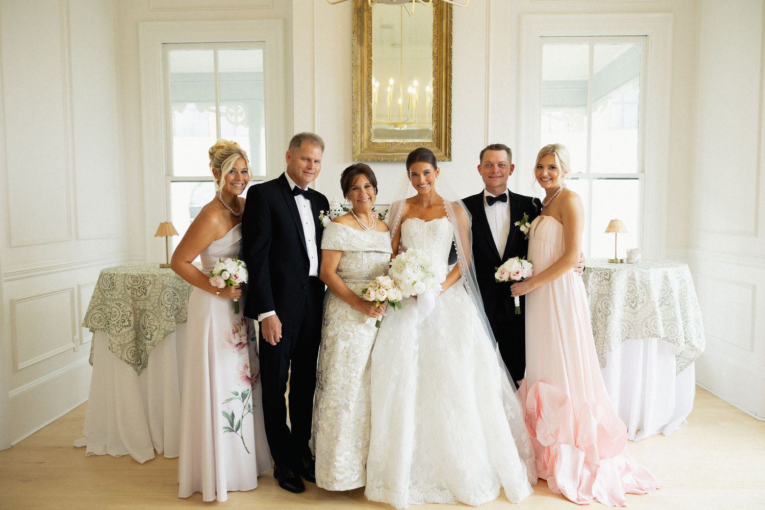 Wedding party poses elegantly in white room, featuring bride in lace gown, groom in tuxedo, and bridesmaids in pink.