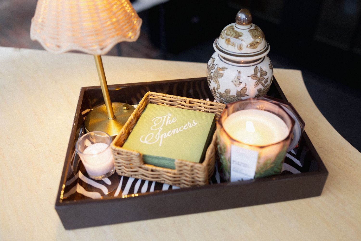 Elegant decorative tray with candles, a wicker basket, green book, ornate jar, and small lamp on a neutral surface.