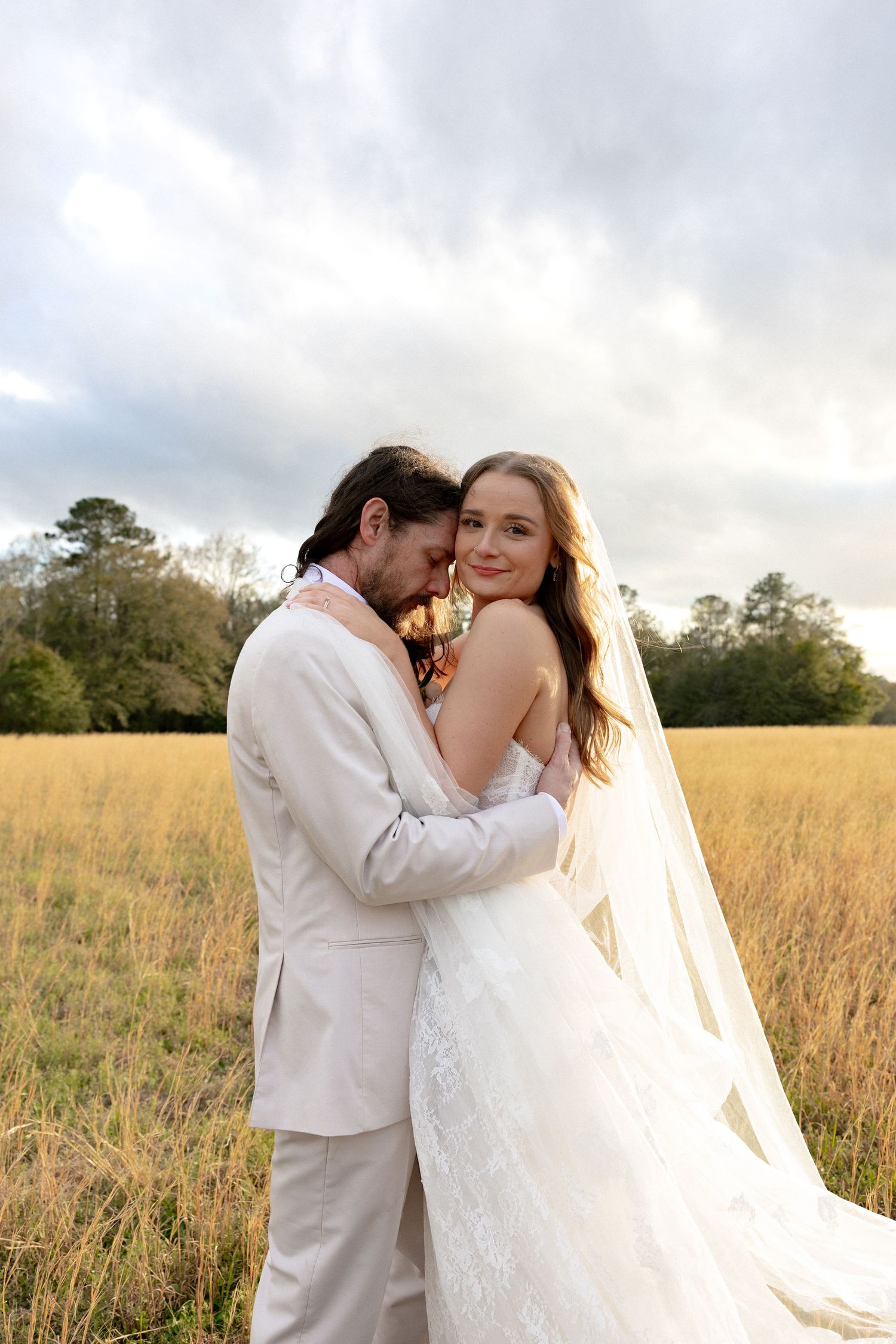 A bride and groom embrace romantically in a golden field, with the bride's veil flowing behind her under a cloudy sky.