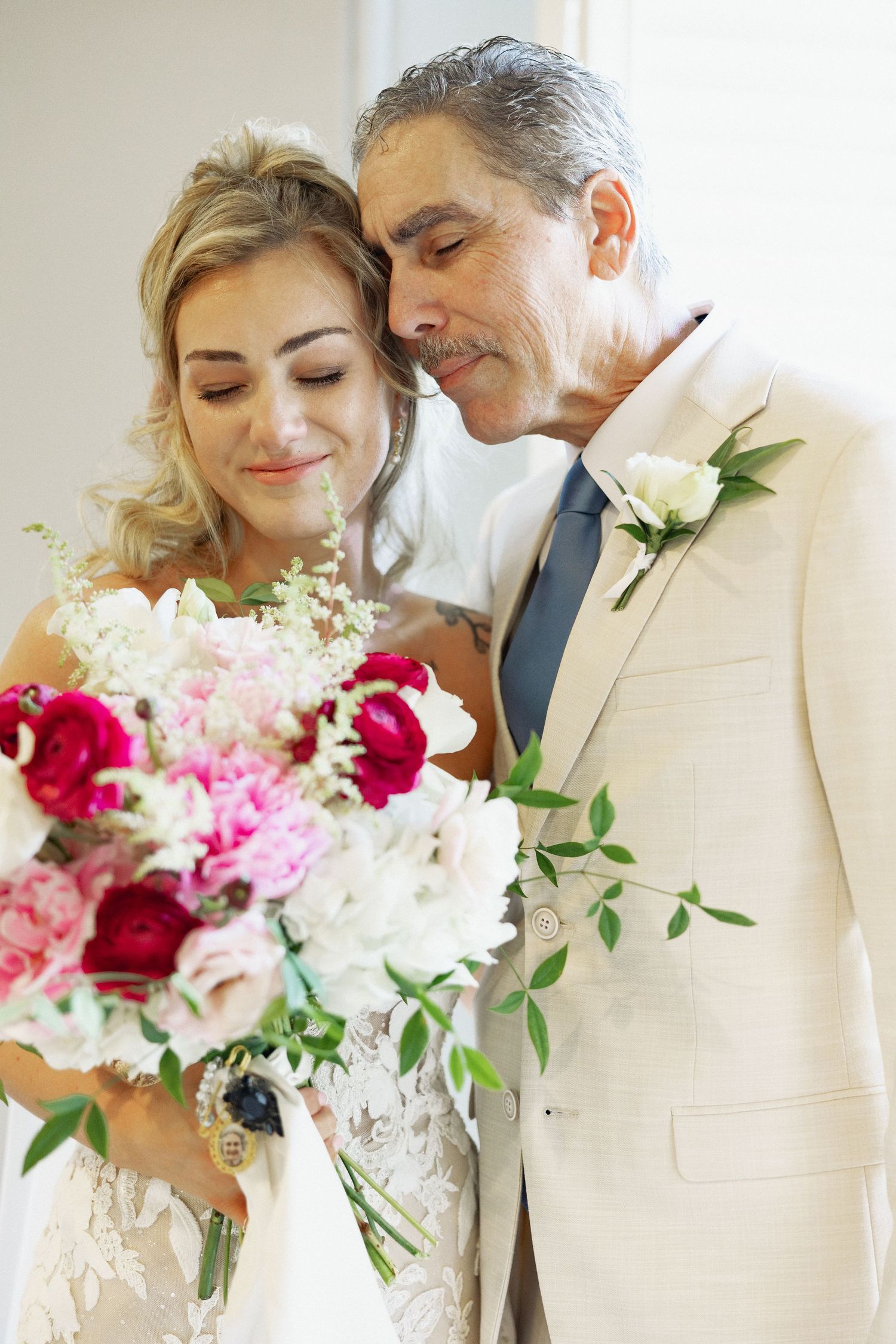 Bride and older gentleman in beige suit share tender moment holding colorful pink and red wedding bouquet.