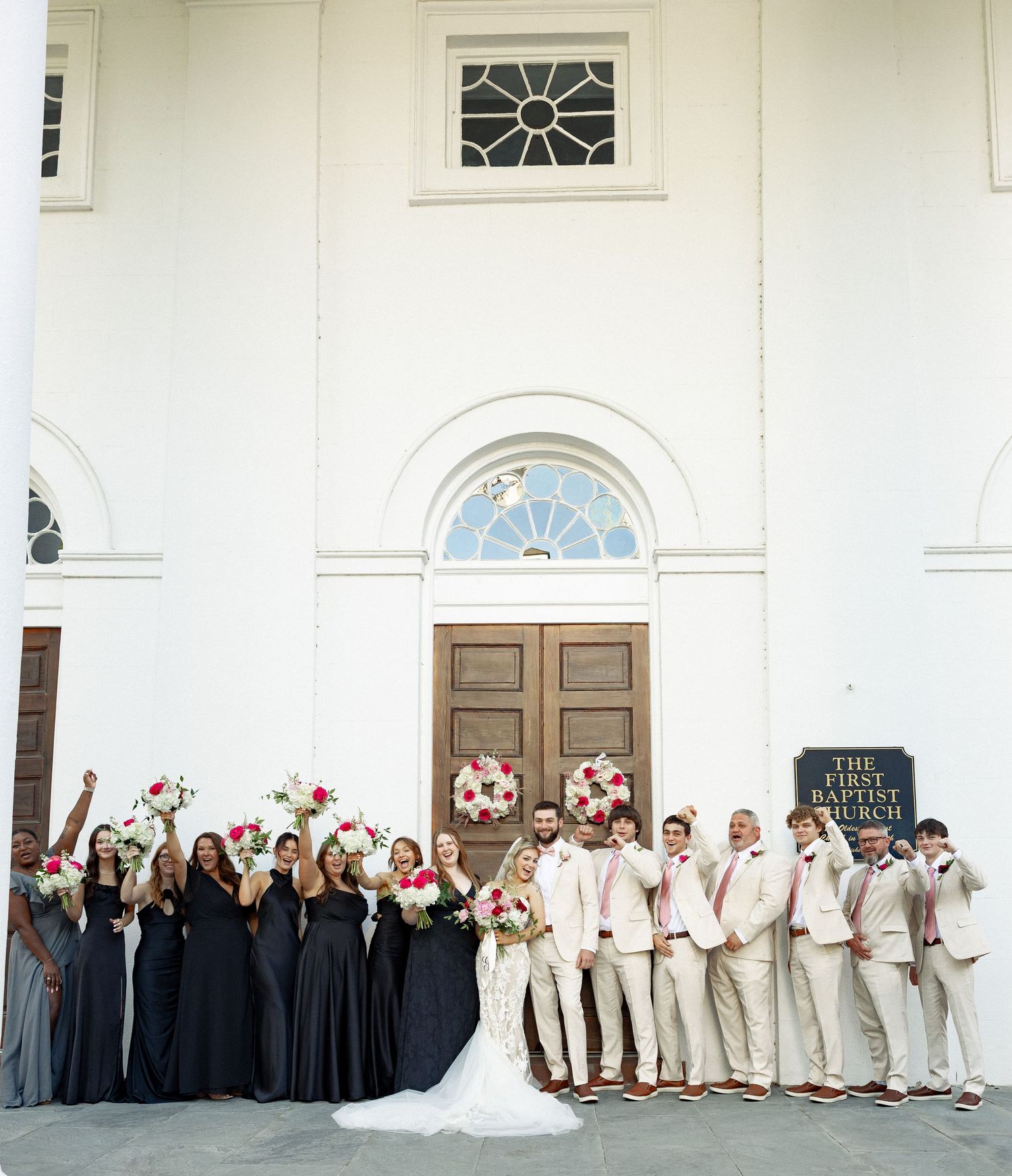 Wedding party celebrating outside First Baptist Church with bride in lace gown and bridesmaids in navy dresses.