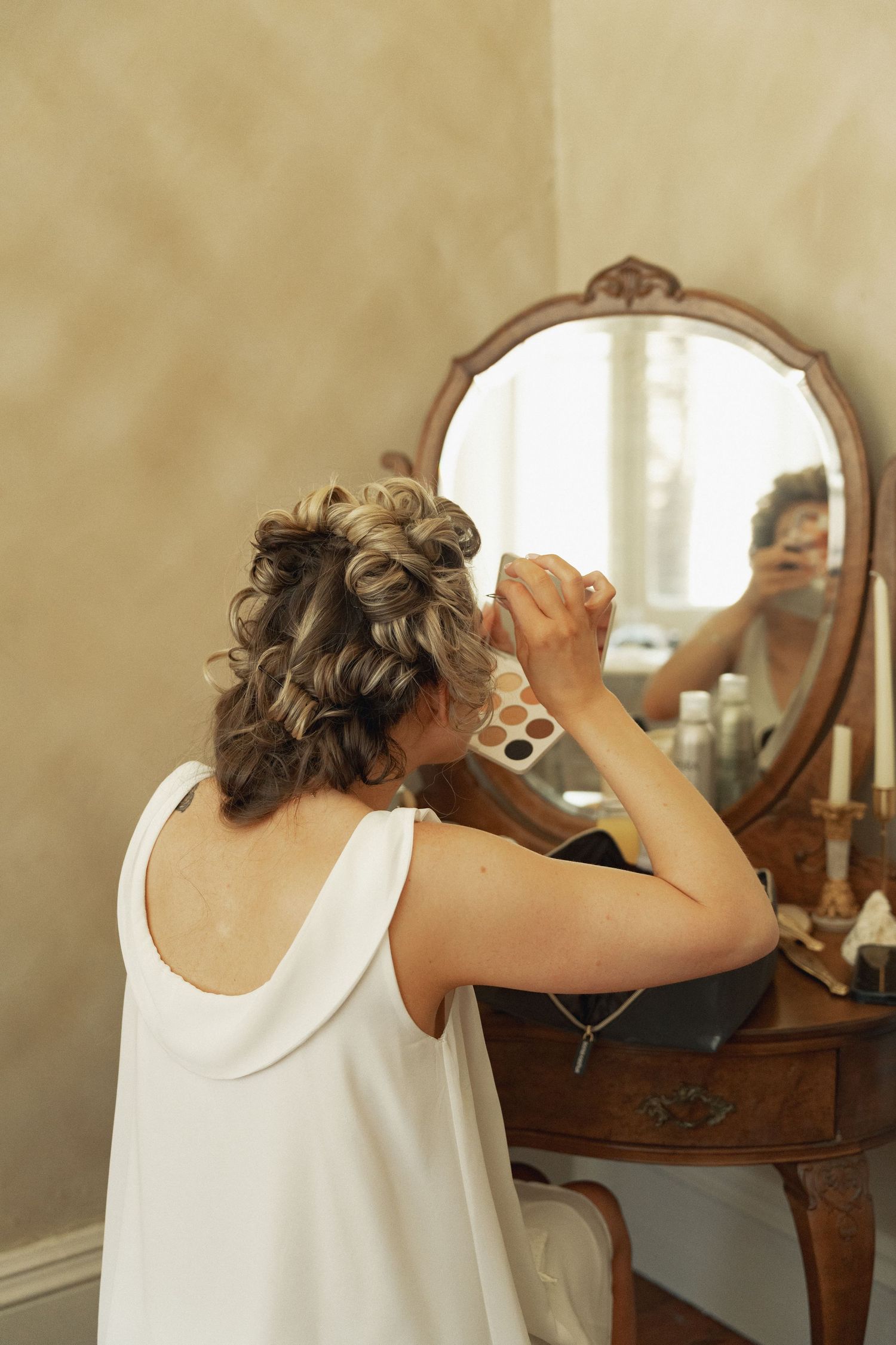 Bride with elegant updo applies makeup at vintage vanity mirror during wedding morning preparations.