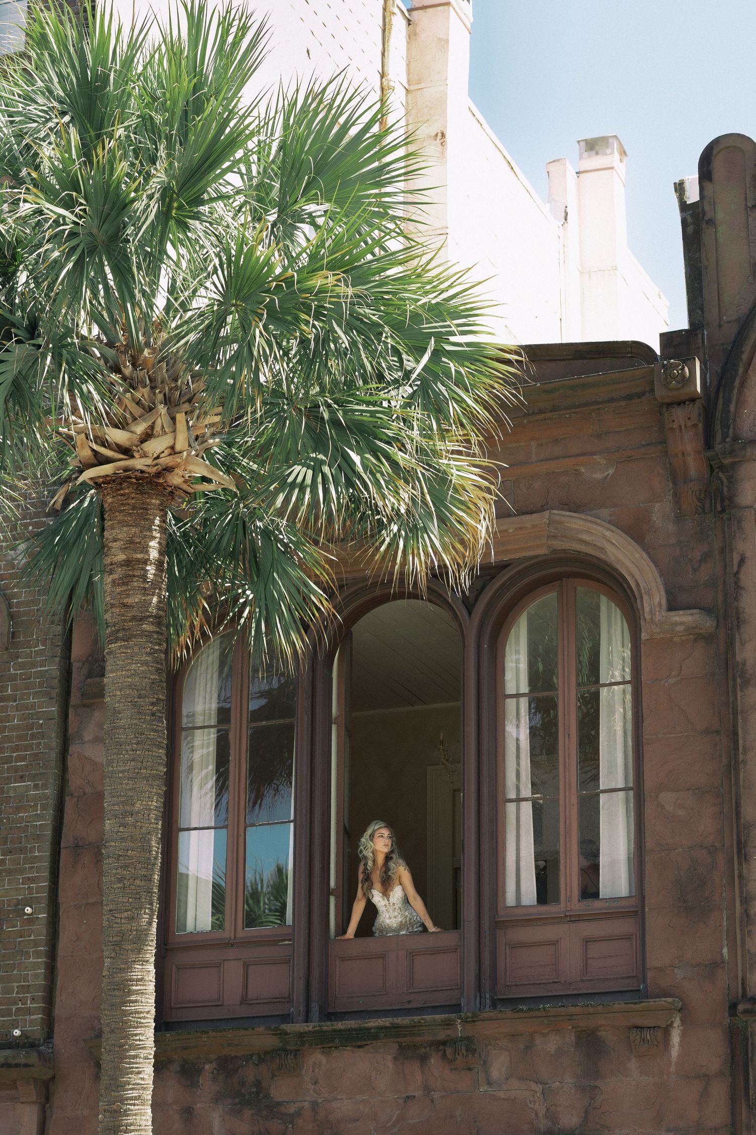 Bride in lace gown sits in arched window of historic brick building with palm tree in Charleston, SC.