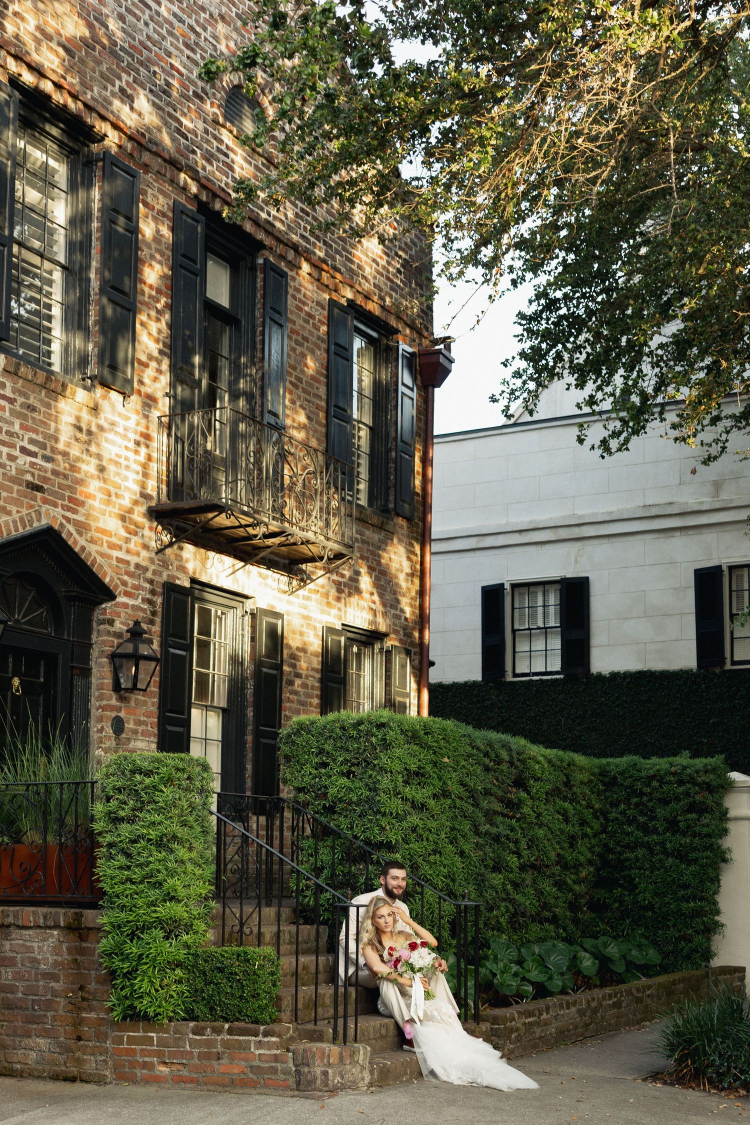 Bride and groom posing on steps of historic Charleston brick building surrounded by lush greenery and black shuttered windows.
