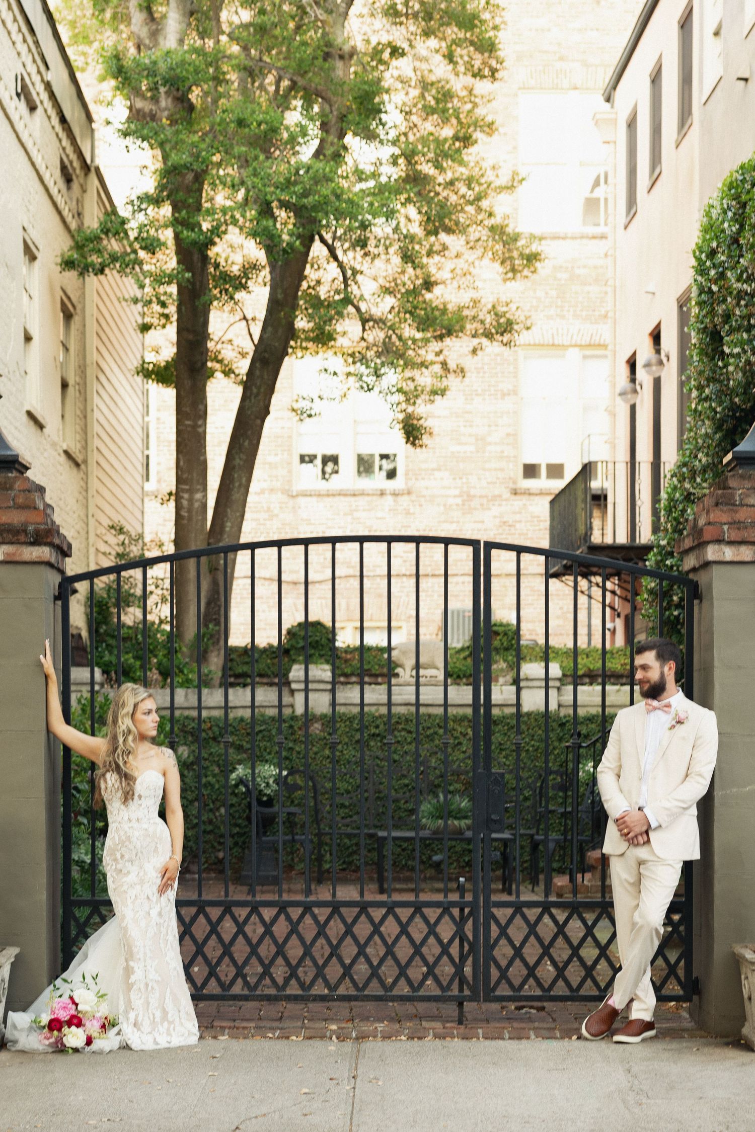 Bride in lace gown and groom in beige suit pose on either side of an ornate black iron garden gate.