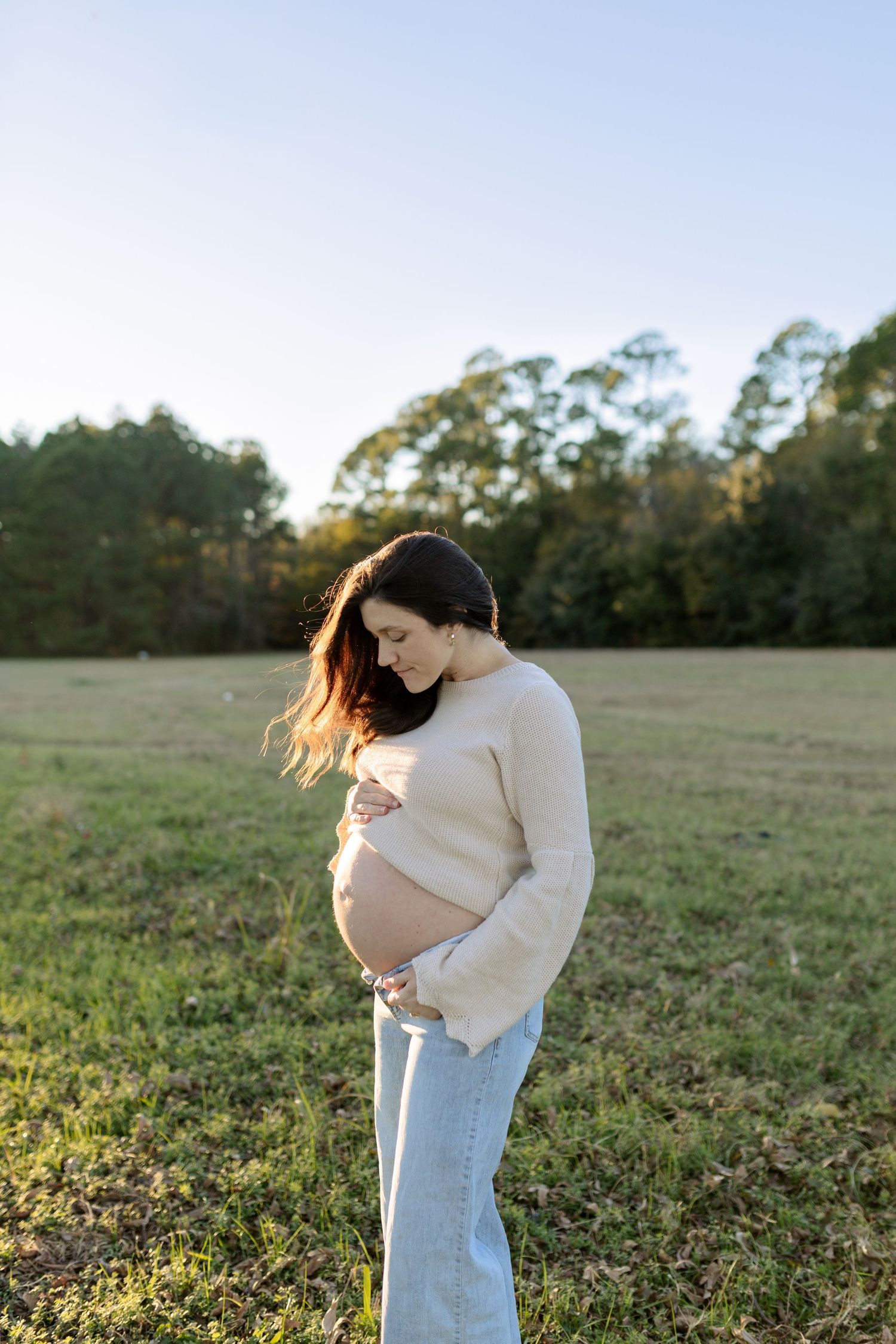 Pregnant woman in beige sweater and jeans holding baby bump in open field at sunset.