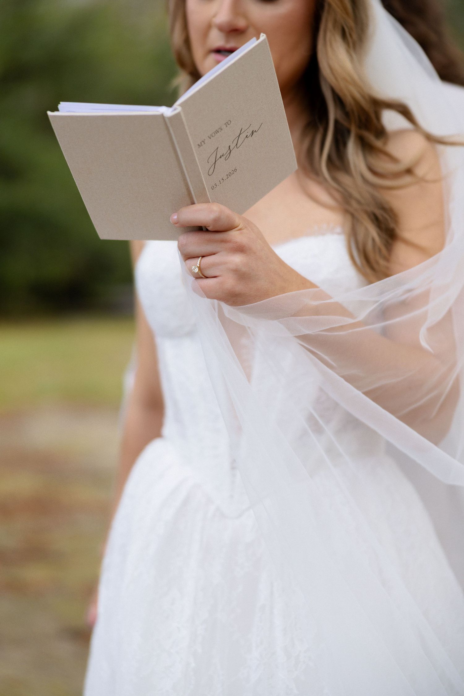 Close-up of bride in strapless lace gown reading personalized vow book with ring on finger.