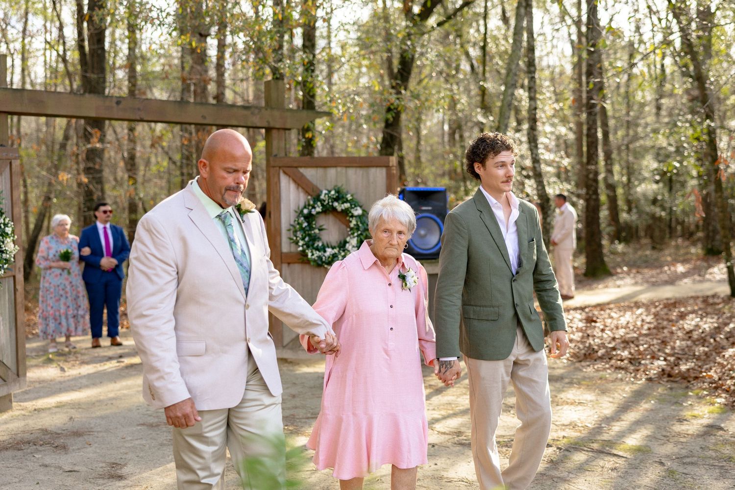 Groom in tan suit escorting elderly woman in pink dress down the aisle at an outdoor woodland wedding.