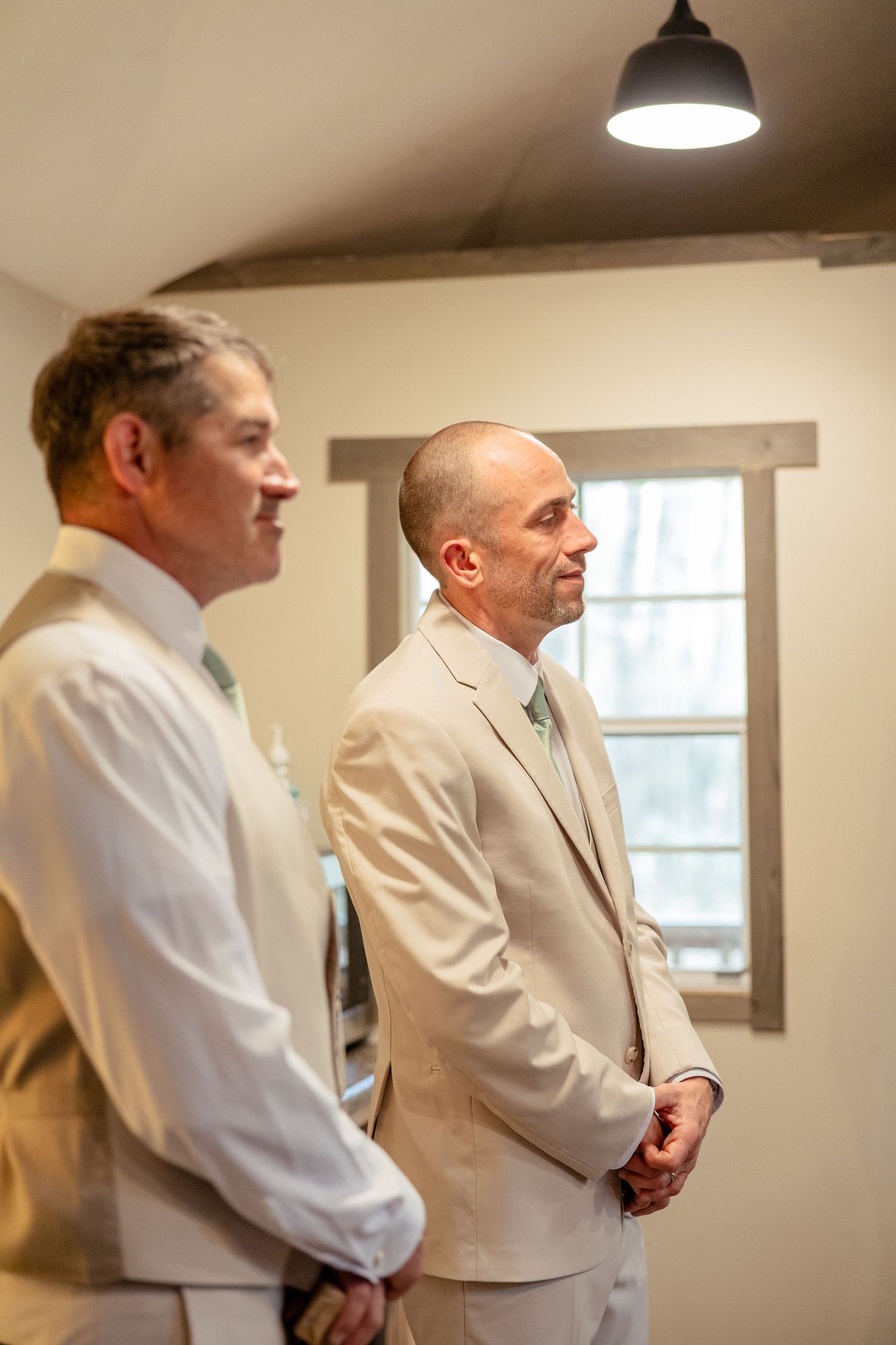Two groomsmen in beige suits stand together under pendant light in a rustic wedding venue room.
