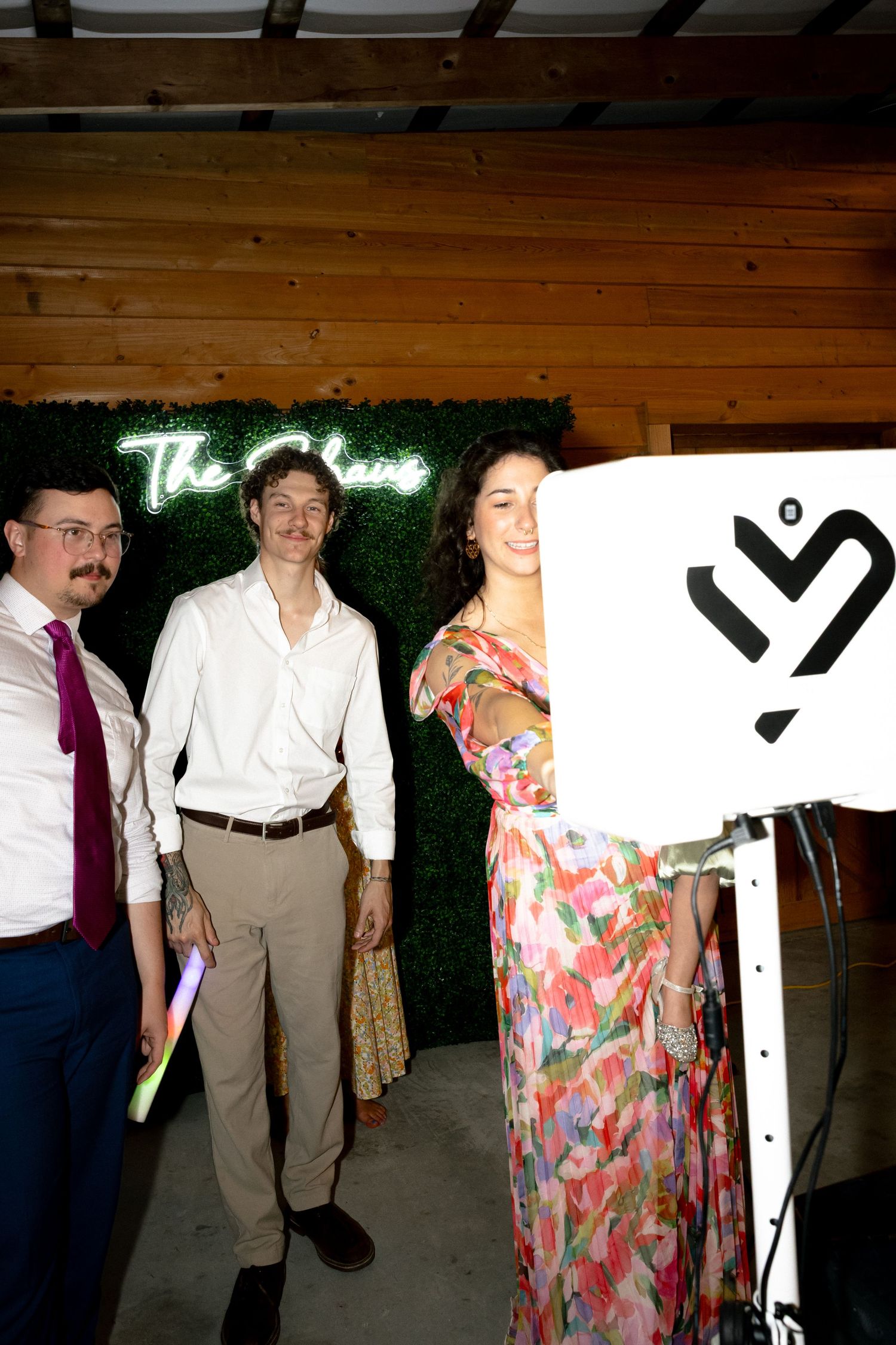 Wedding guests in formal attire pose near neon sign and photo booth at elegant barn reception venue.
