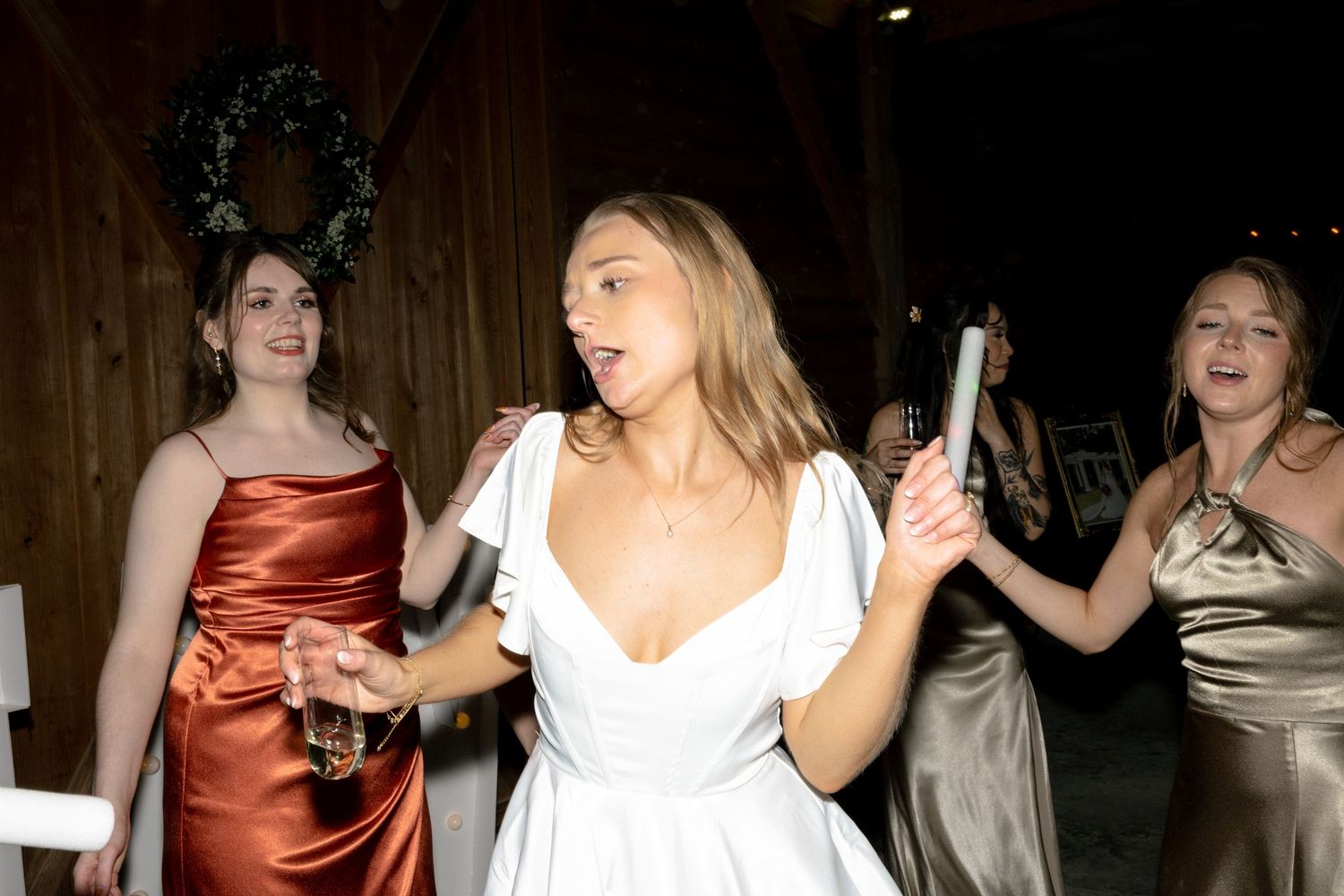 Bride in white dress dances joyfully with bridesmaids in colorful gowns at wedding reception.