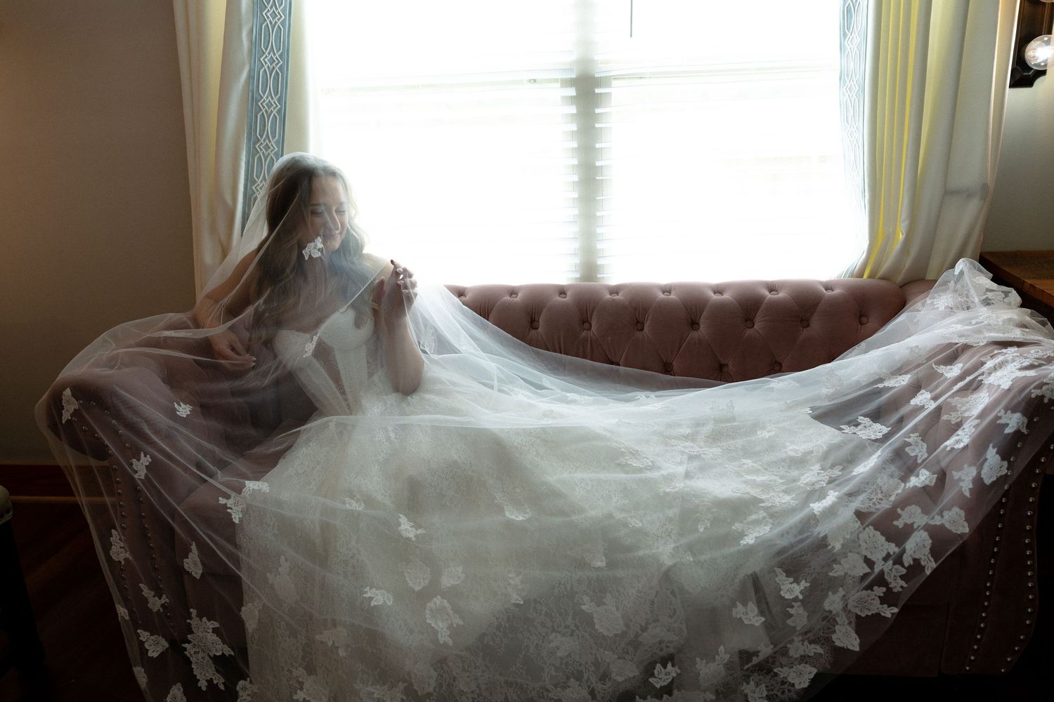 Bride in lace wedding dress with long floral veil spread across pink sofa, gazing out window.