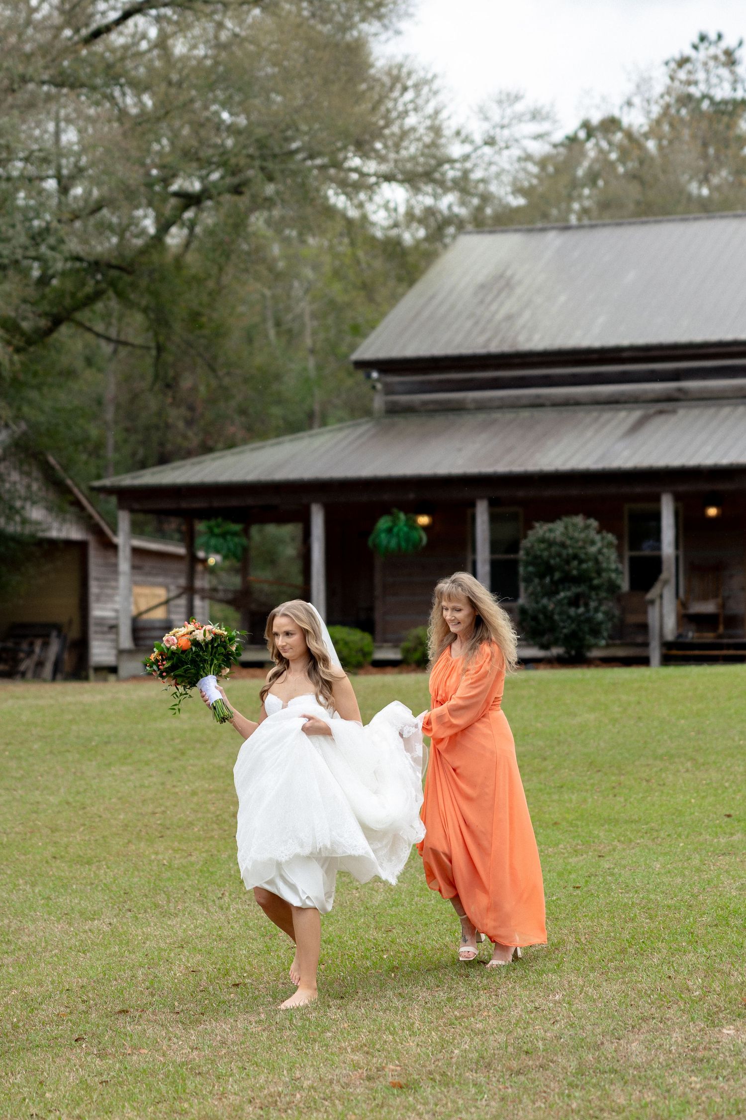 Bride in white gown holding orange bouquet walks barefoot on grass assisted by woman in orange dress.