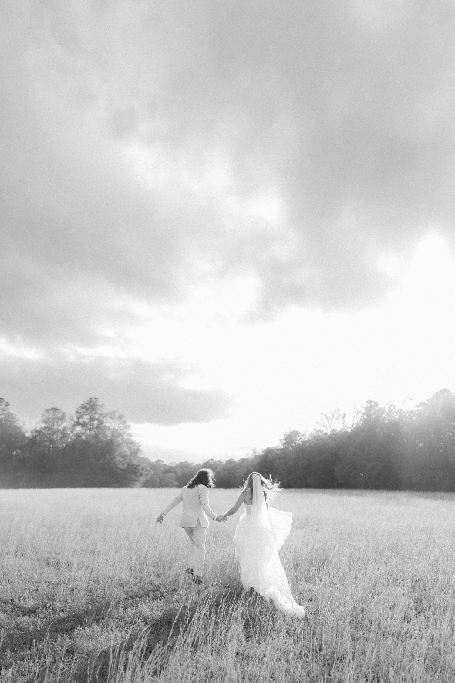 Black and white photo of bride and groom running through a sunlit field on their wedding day.