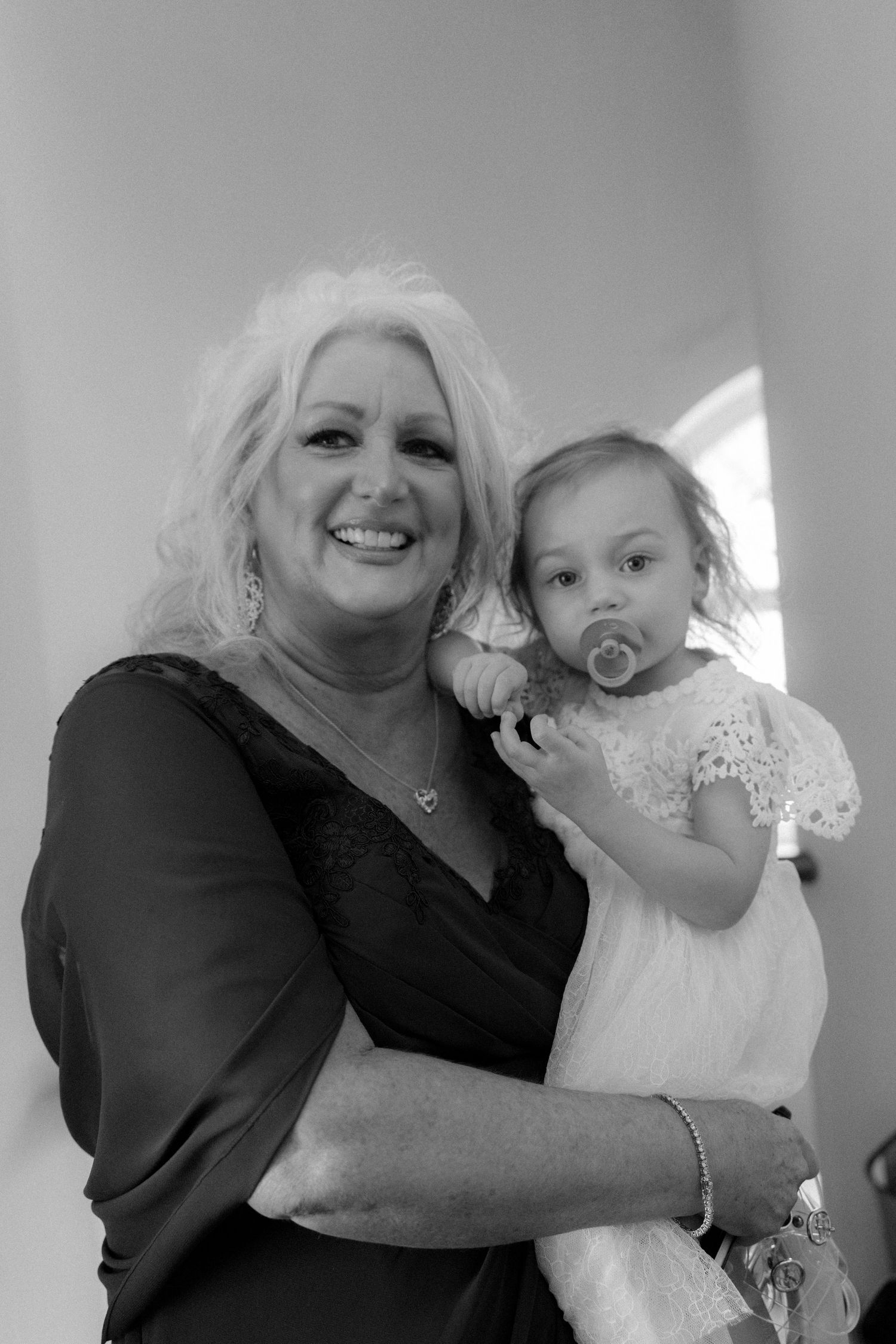 Smiling woman with blonde curly hair holds a baby girl with pacifier in a black and white portrait.