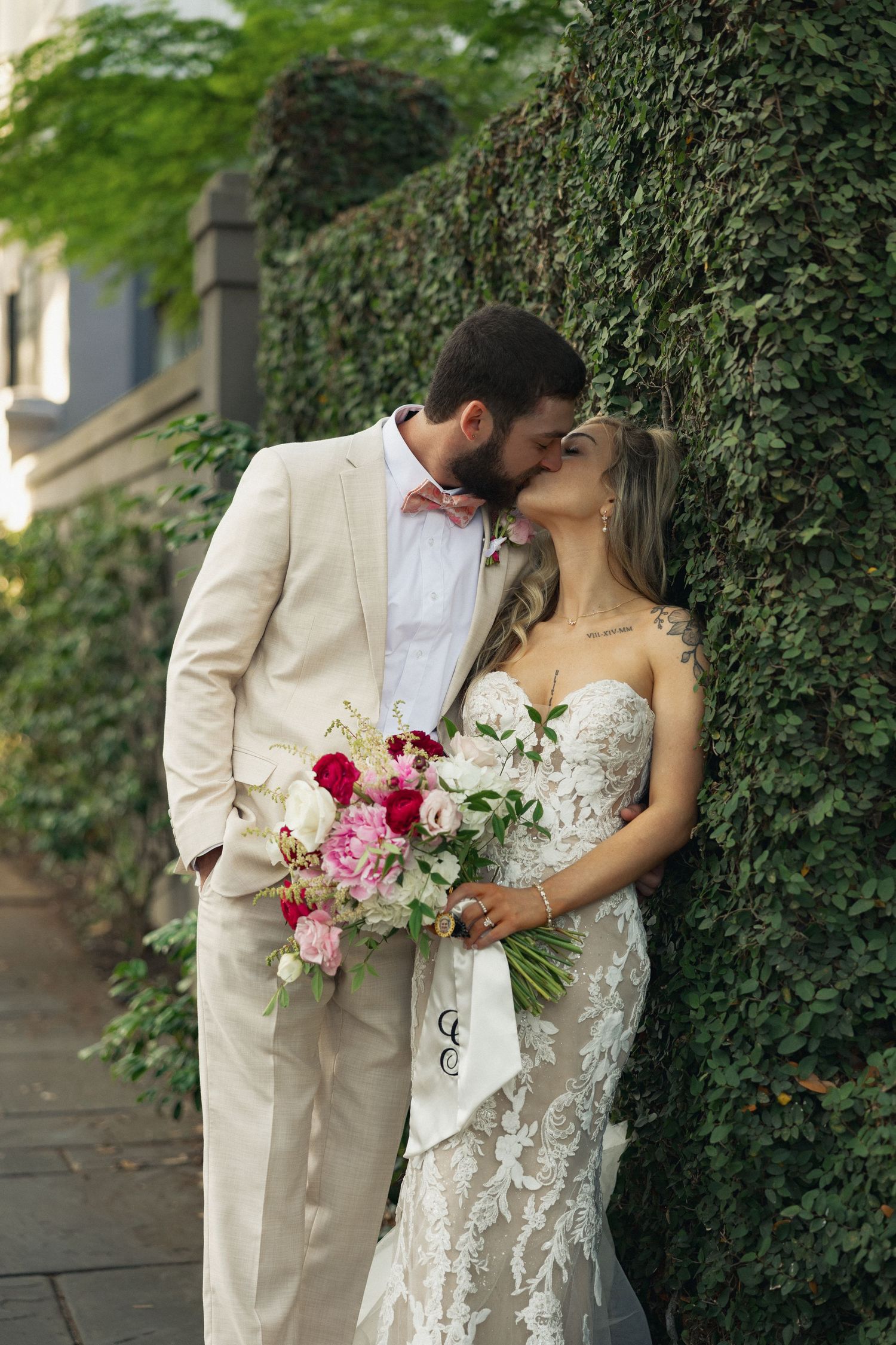 Bride and groom kissing against ivy-covered wall, Charleston wedding, lace dress, tan suit, pink bow tie, floral bouquet.
