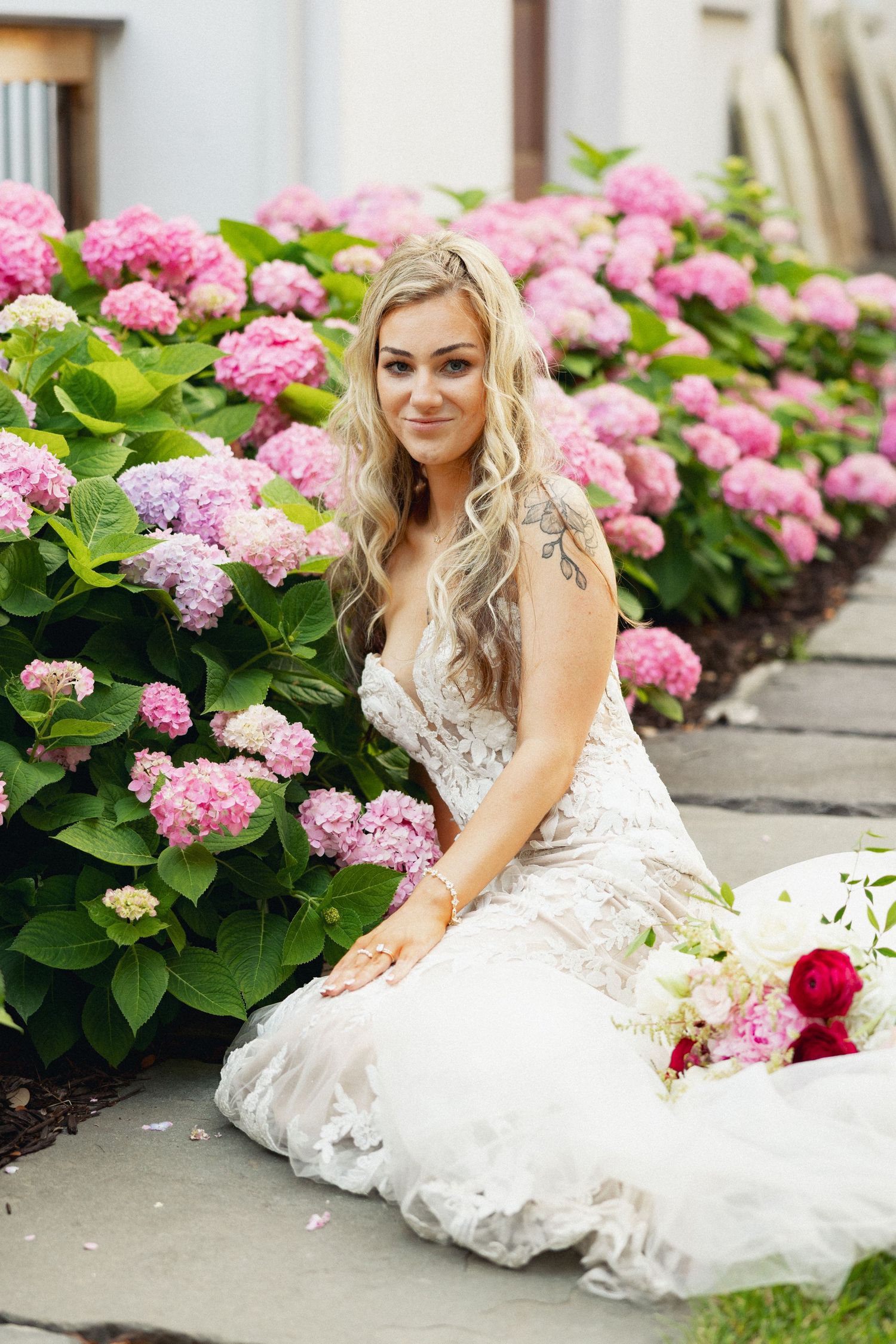 Blonde bride in lace wedding gown sitting beside pink hydrangea bushes holding a bouquet of white and red flowers.