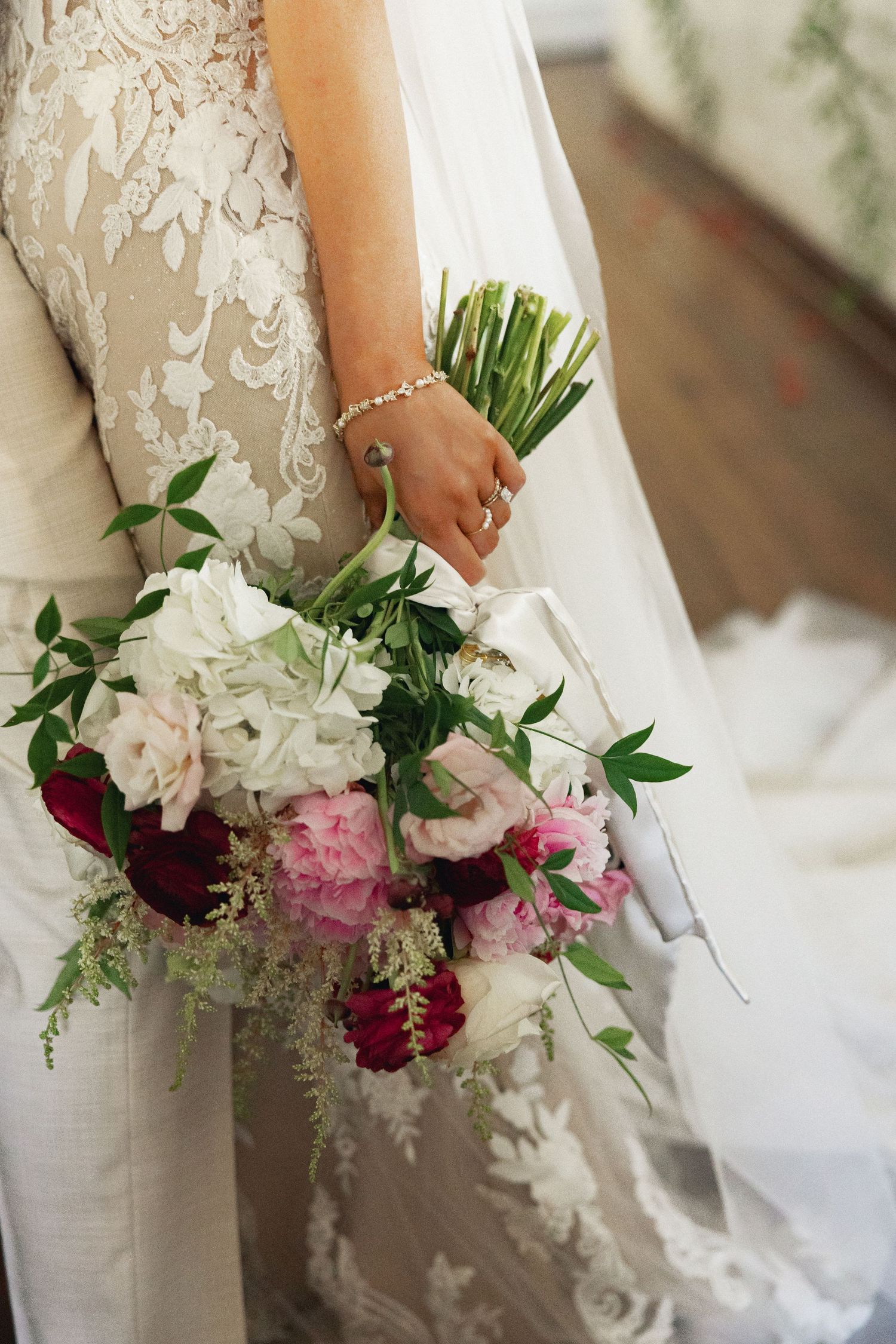 Bride in lace wedding gown holding a lush bridal bouquet of white, pink, and burgundy flowers with greenery.