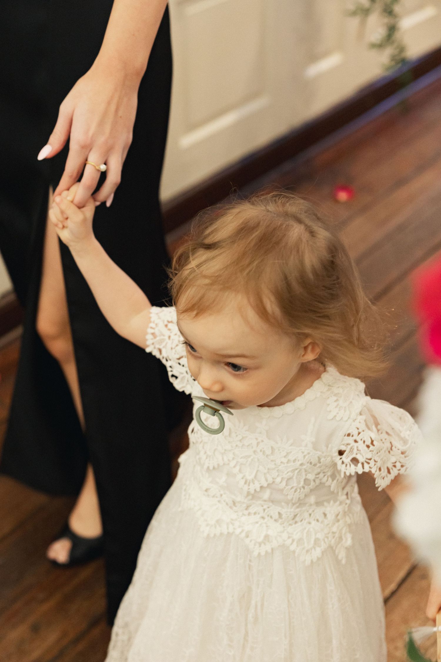 Adorable toddler flower girl in white lace dress holding an adults hand during wedding ceremony.