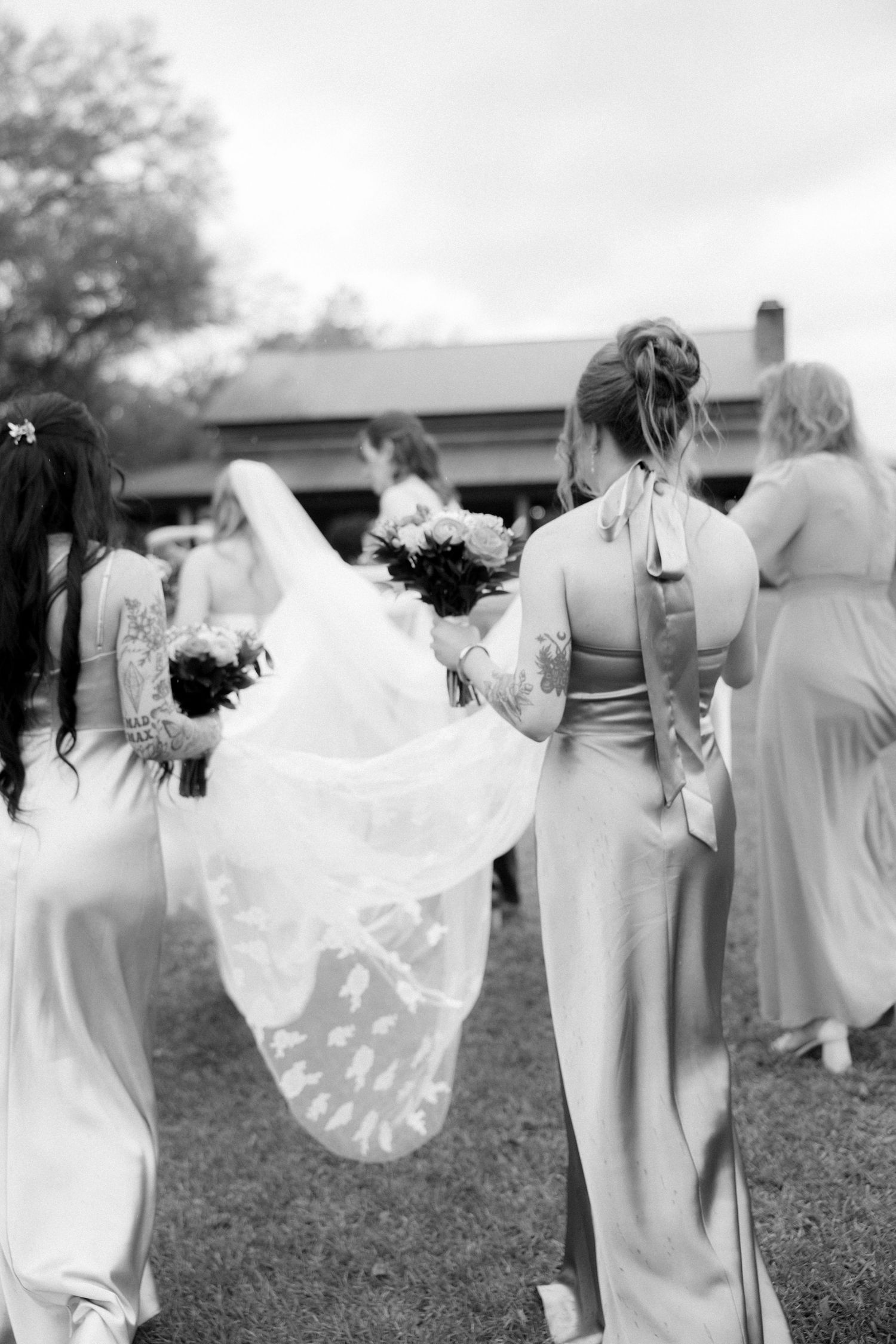 Bridesmaids in elegant gowns carry bride's lace veil and train walking toward rustic venue in black and white.