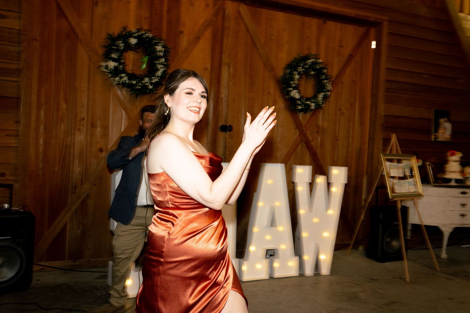 Woman in rust satin gown clapping at elegant barn wedding reception with marquee letter display.