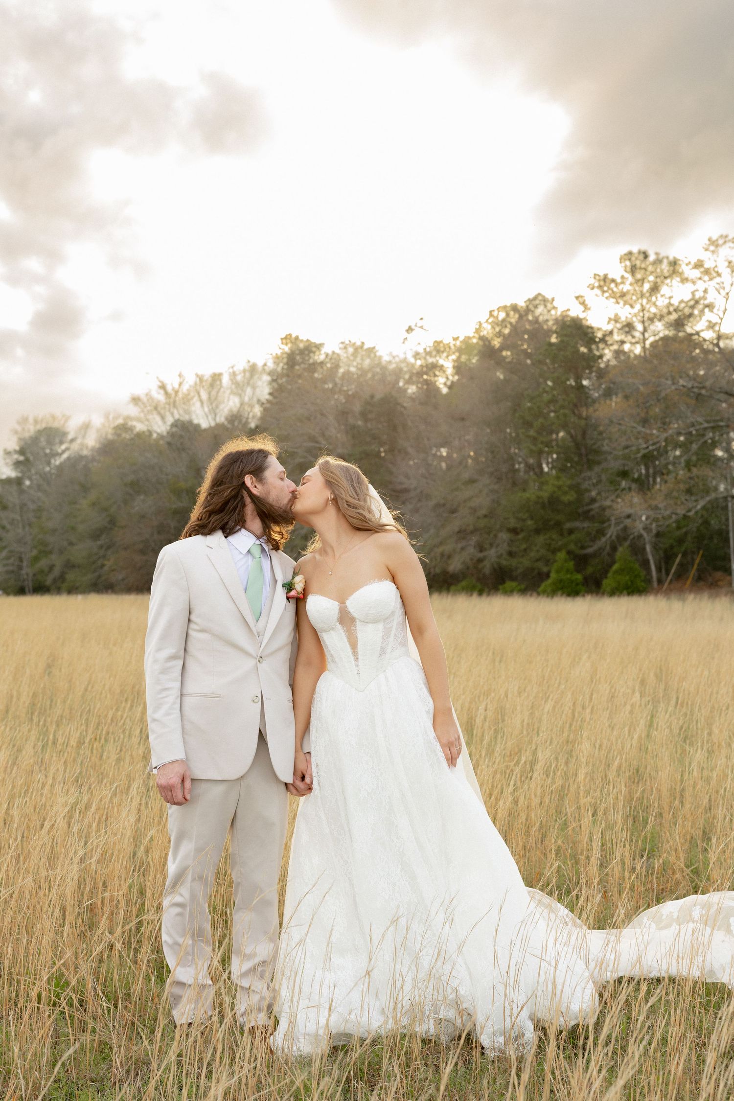 Bride and groom sharing a romantic kiss in a golden field at sunset, groom in beige suit, bride in white strapless gown.