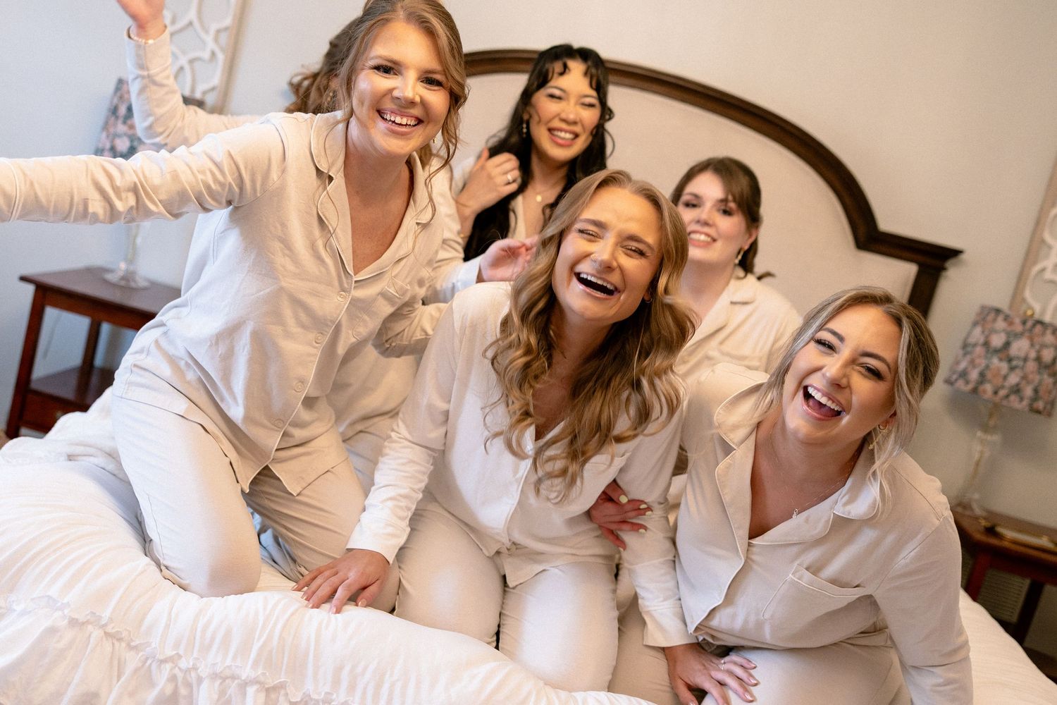 Four bridesmaids in white pajamas laughing and posing on a bed together.