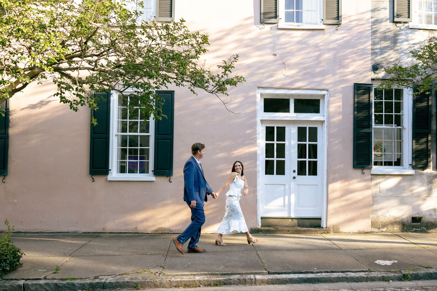 Couple walking together in front of charming pink building with green shutters.