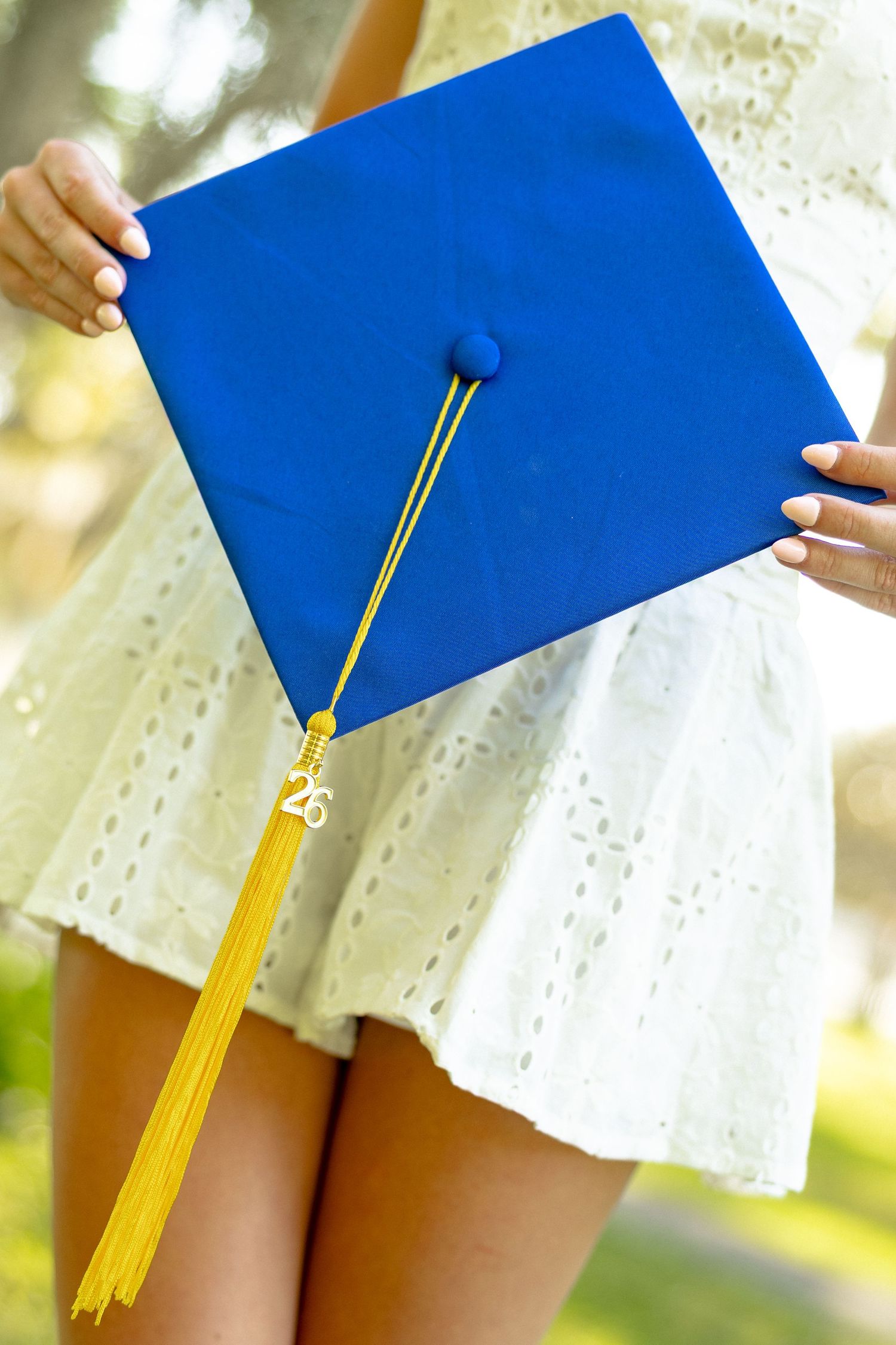 Blue graduation cap with gold tassel held by graduate in white lace dress outdoors.