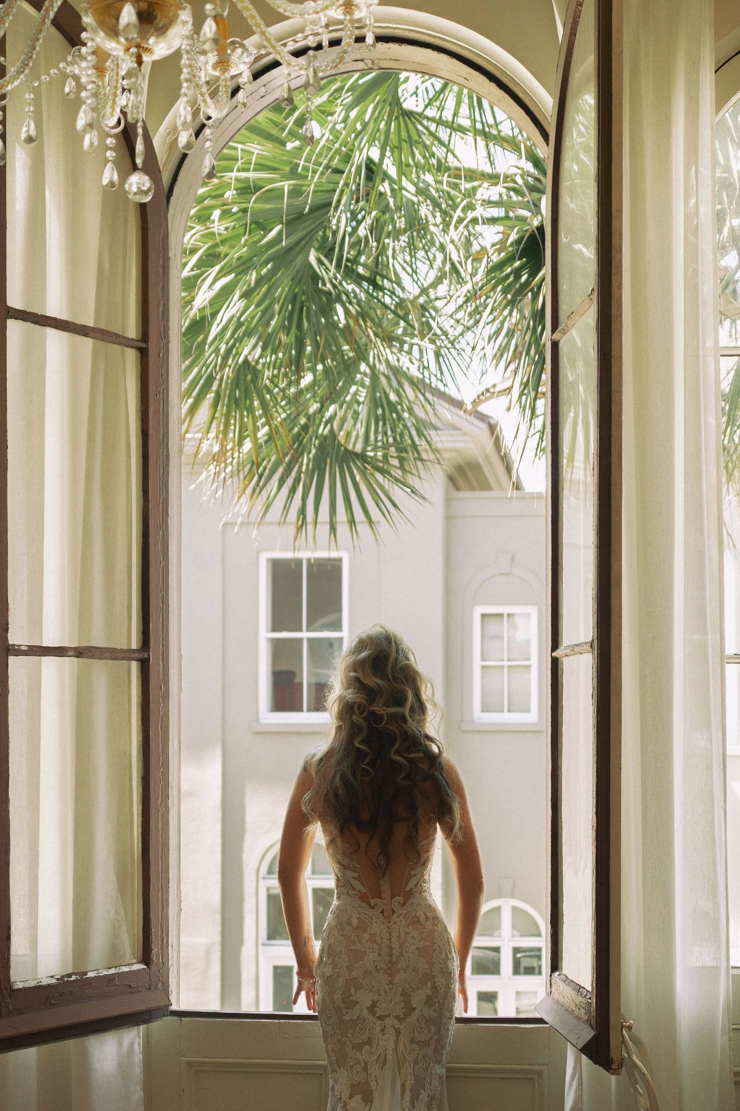 Bride in lace wedding dress stands in open French doors overlooking a palm tree and white building.