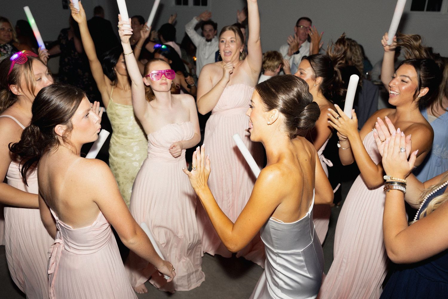 Young women in pink bridesmaid dresses dancing joyfully at a wedding reception party with glow sticks.