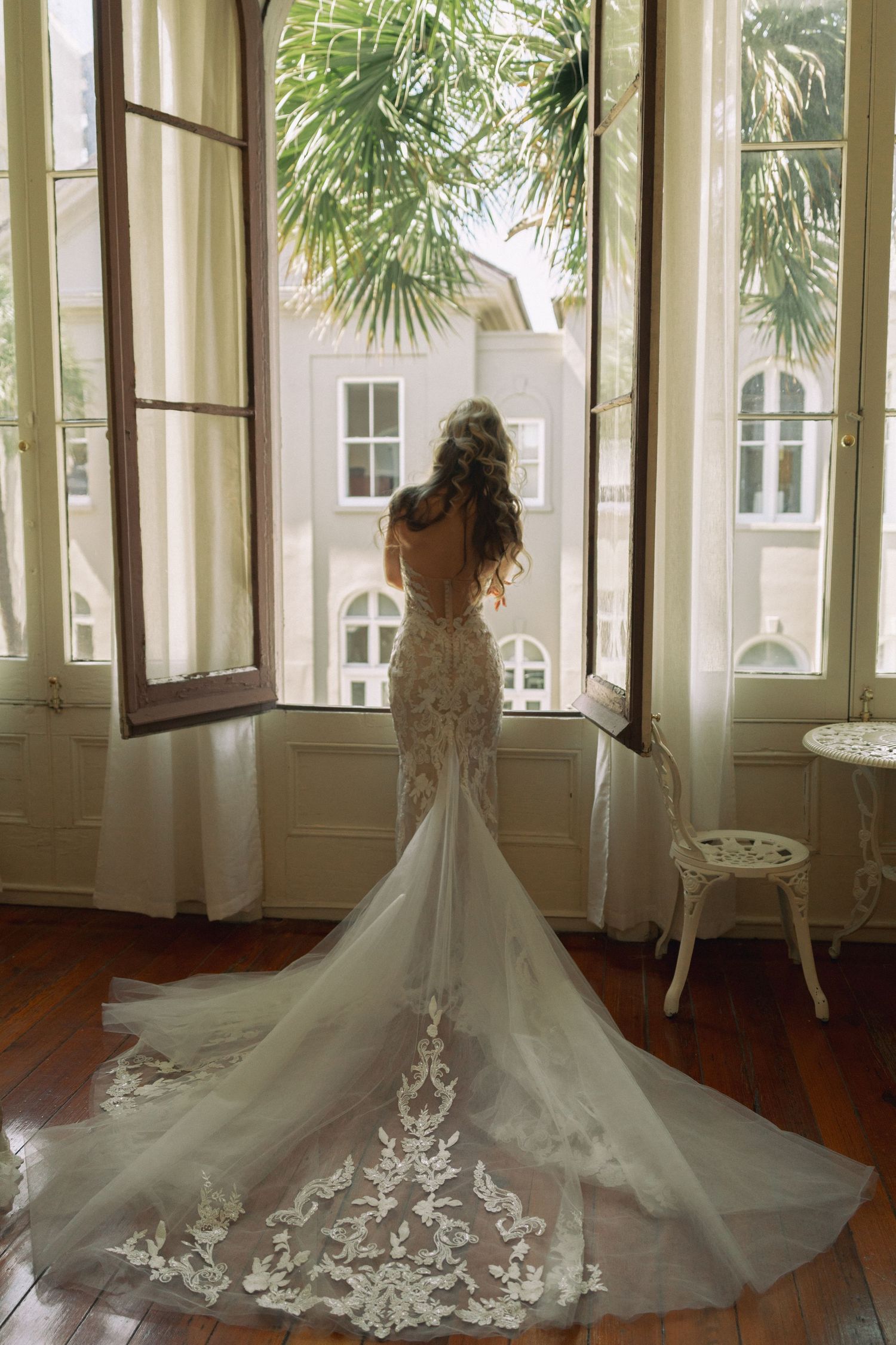 Bride in elegant lace mermaid wedding gown with long train stands by open French doors overlooking tropical garden.
