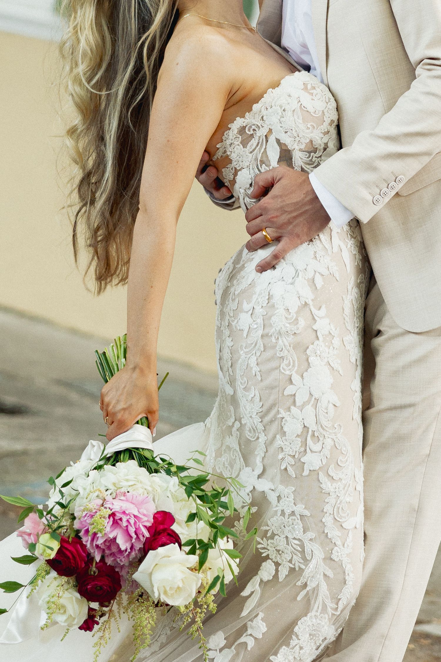 Close-up of bride in intricate lace wedding gown holding pink and white floral bouquet, groom in tan suit embracing her.