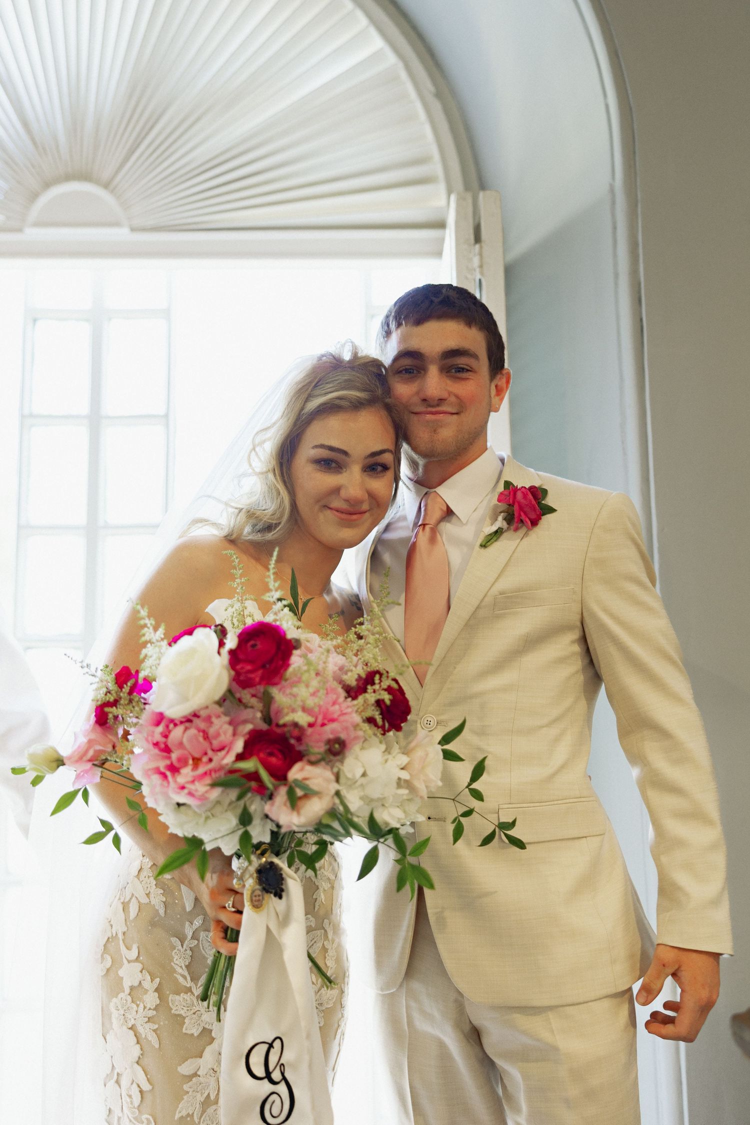 Bride and groom pose together, she holds pink floral bouquet, he wears tan suit with pink boutonniere, near arched window.