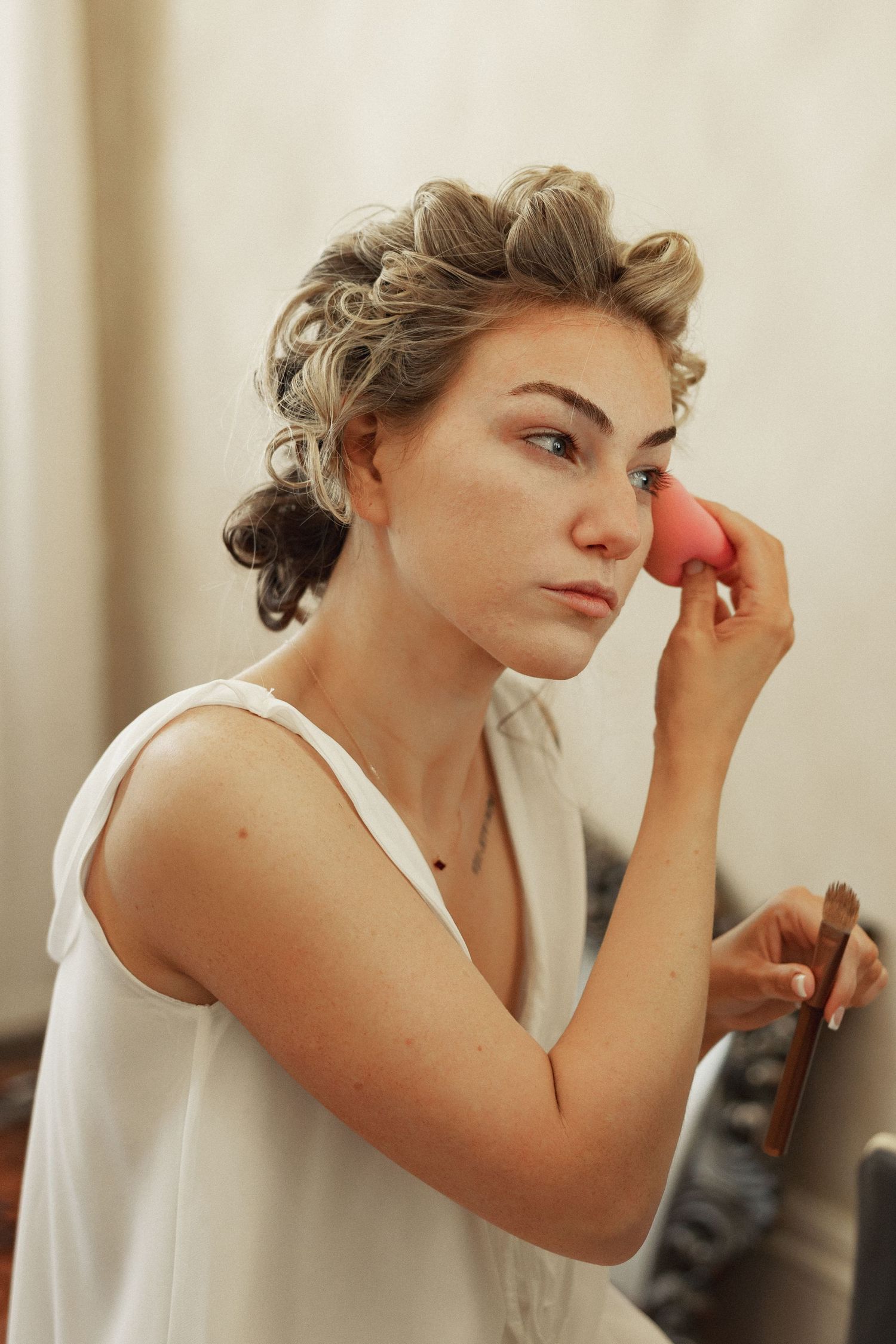 A young bride applies pink makeup sponge to her cheek while getting ready for her wedding day.