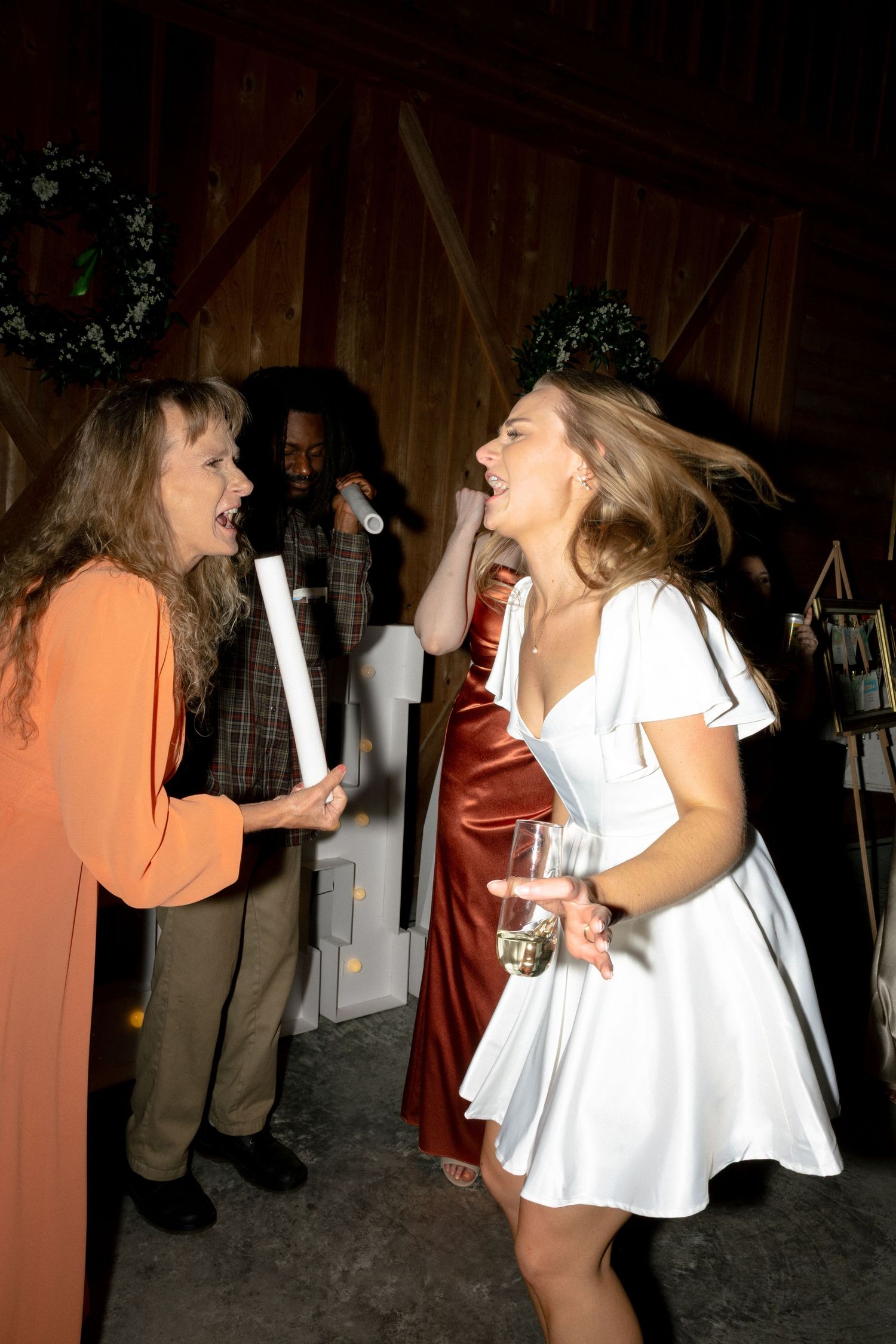 Bride in white dress laughs with a woman in orange at a lively barn wedding reception.