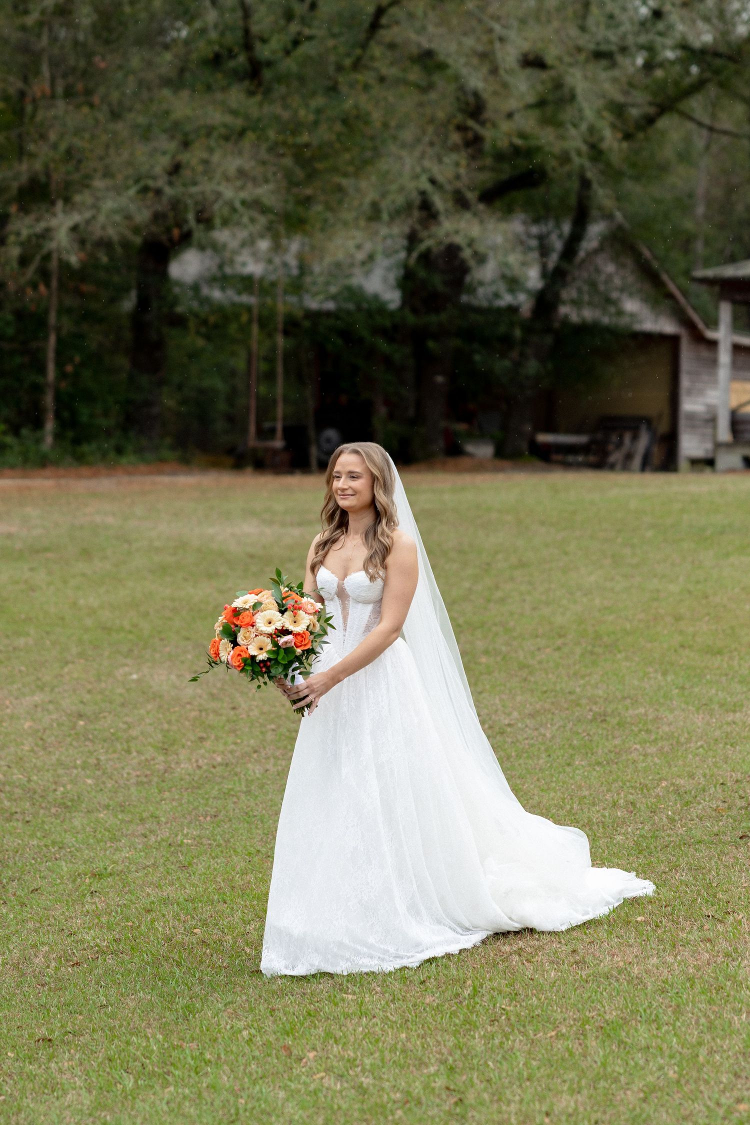 Elegant bride in white ball gown with long veil holding orange bouquet on green lawn outdoors.