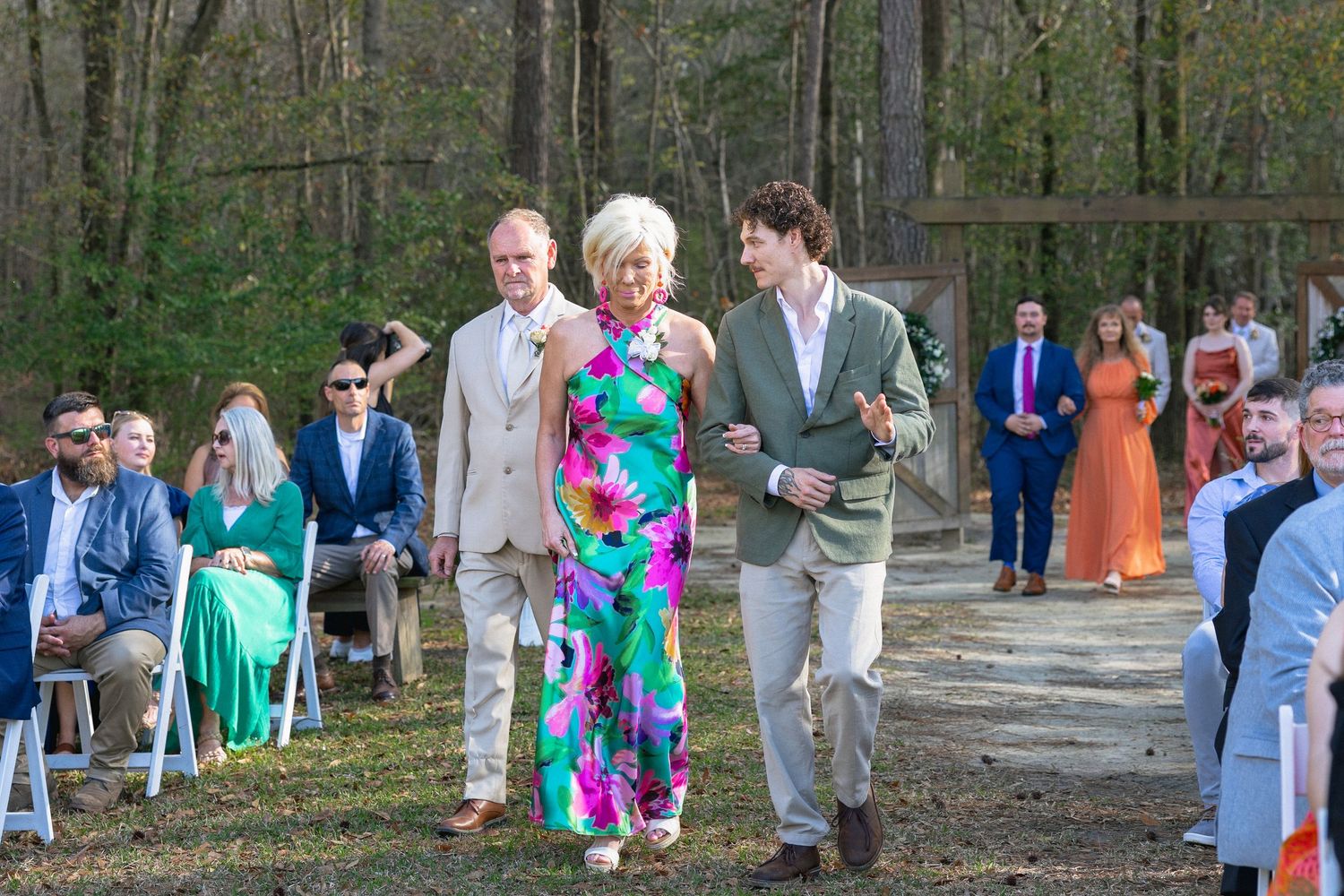 Groom escorting woman in colorful floral jumpsuit down the aisle at an outdoor woodland wedding ceremony.