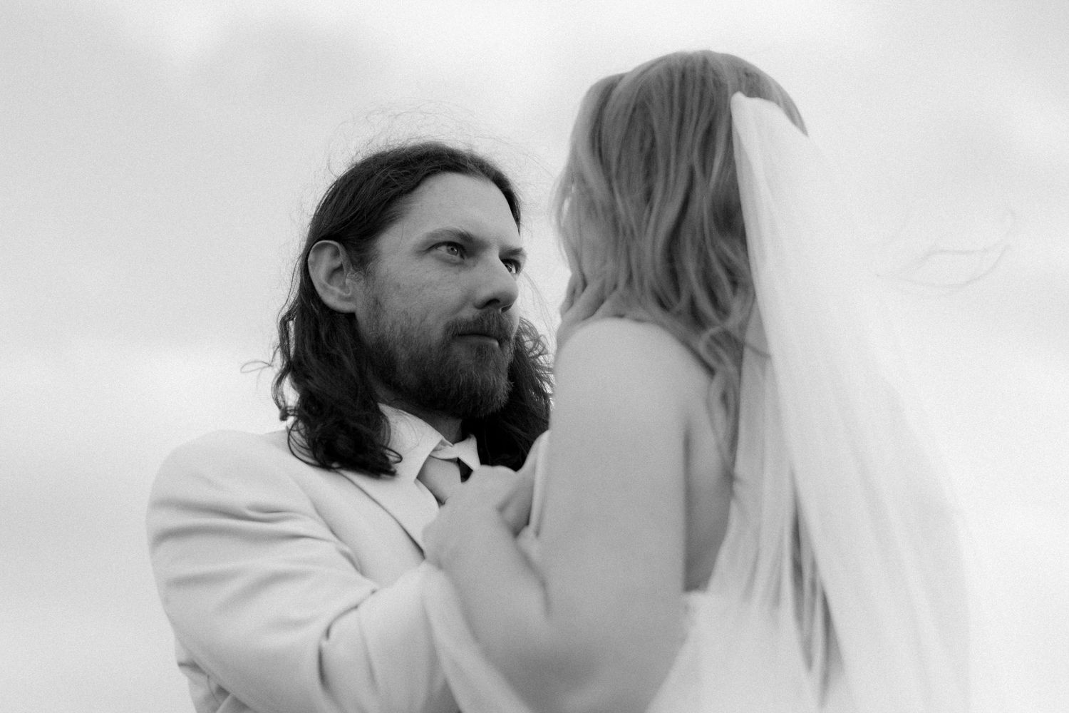 Black and white photo of bearded groom in white suit gazing at blonde bride in veil.